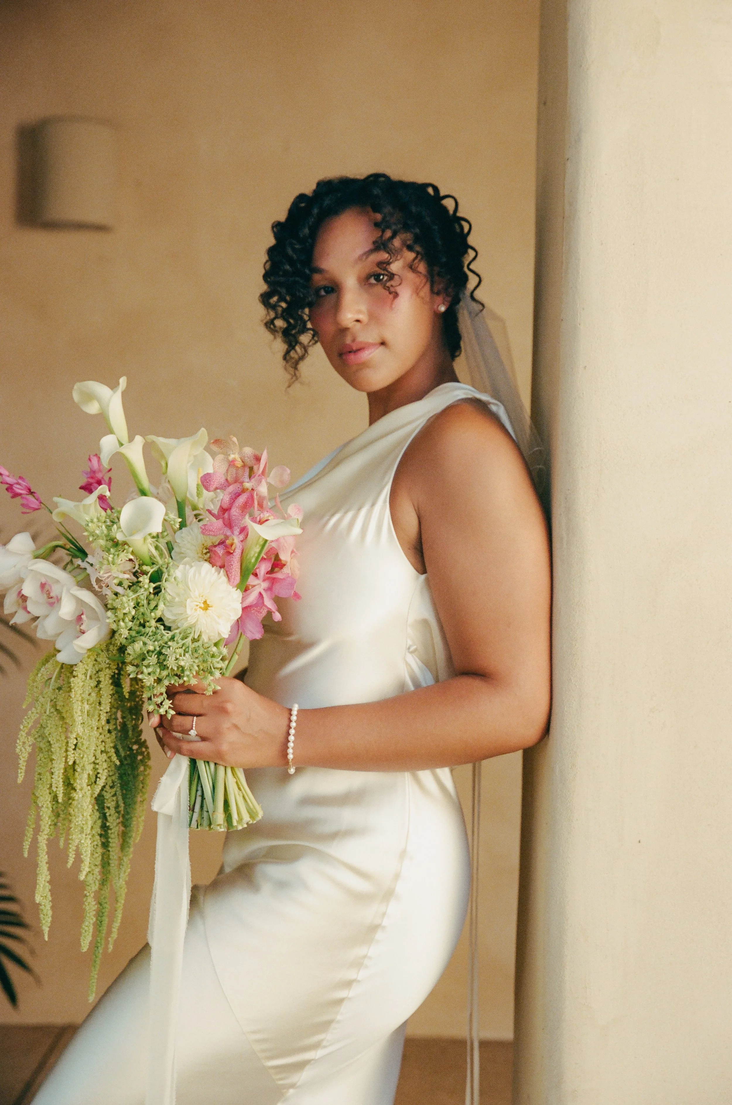 A woman in a white satin sleeveless dress holding a large bouquet of white and pink flowers, standing against a beige wall, looking at the camera with a neutral expression.
