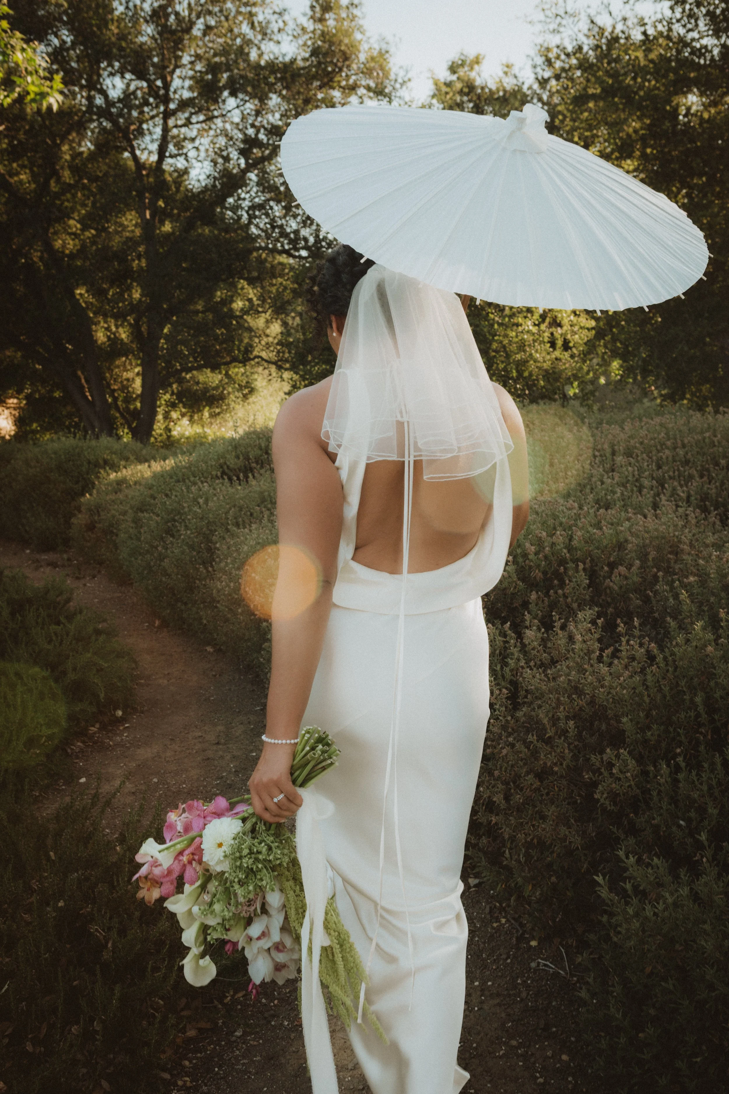 A woman dressed in a white wedding gown, holding a bouquet of flowers, standing outdoors in a garden with trees, holding a white parasol, with her back facing the camera.