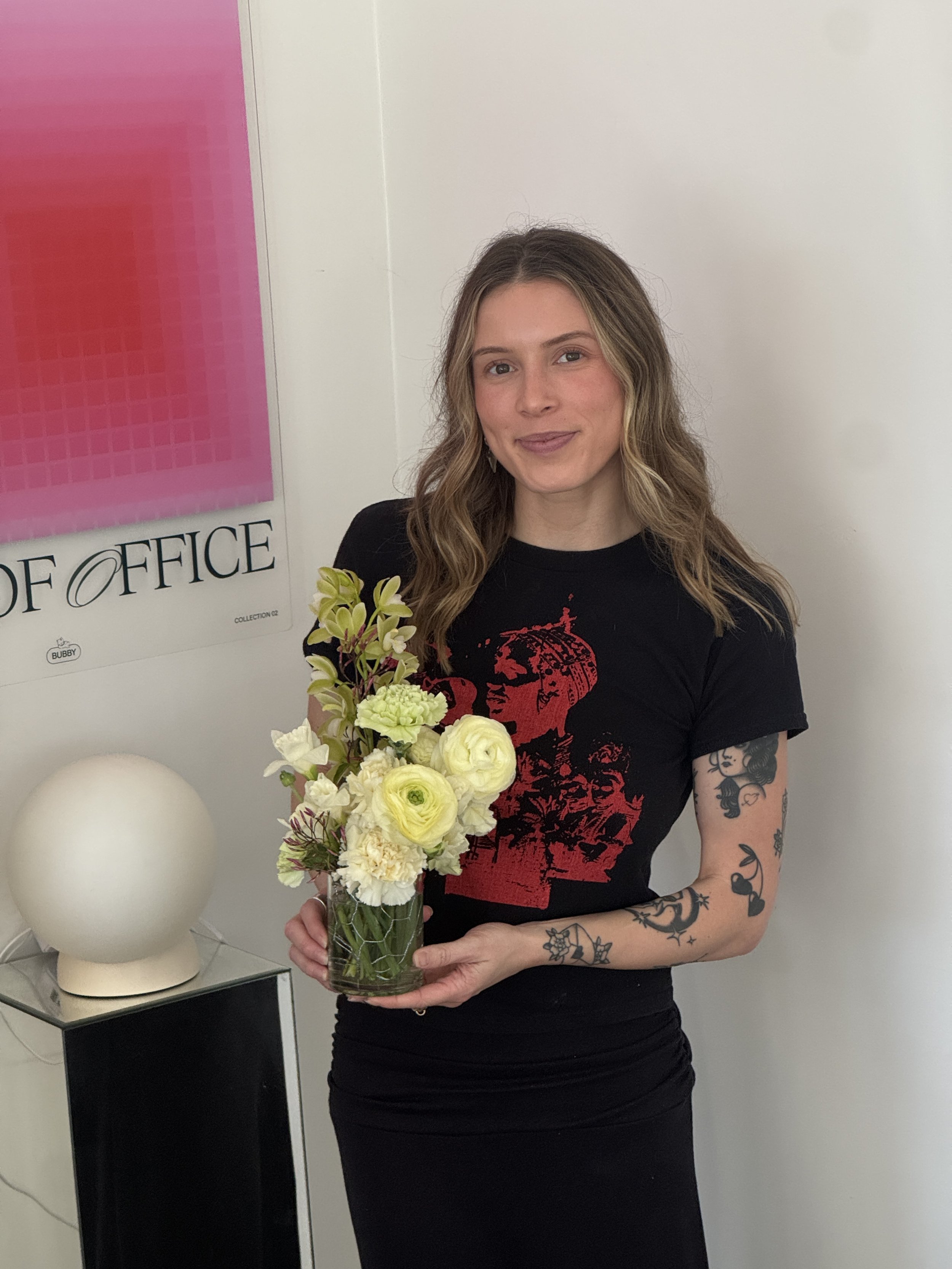A woman with long wavy hair holding a glass vase with white and yellow flowers, standing in a room with white walls, near a pink and white poster on the wall.