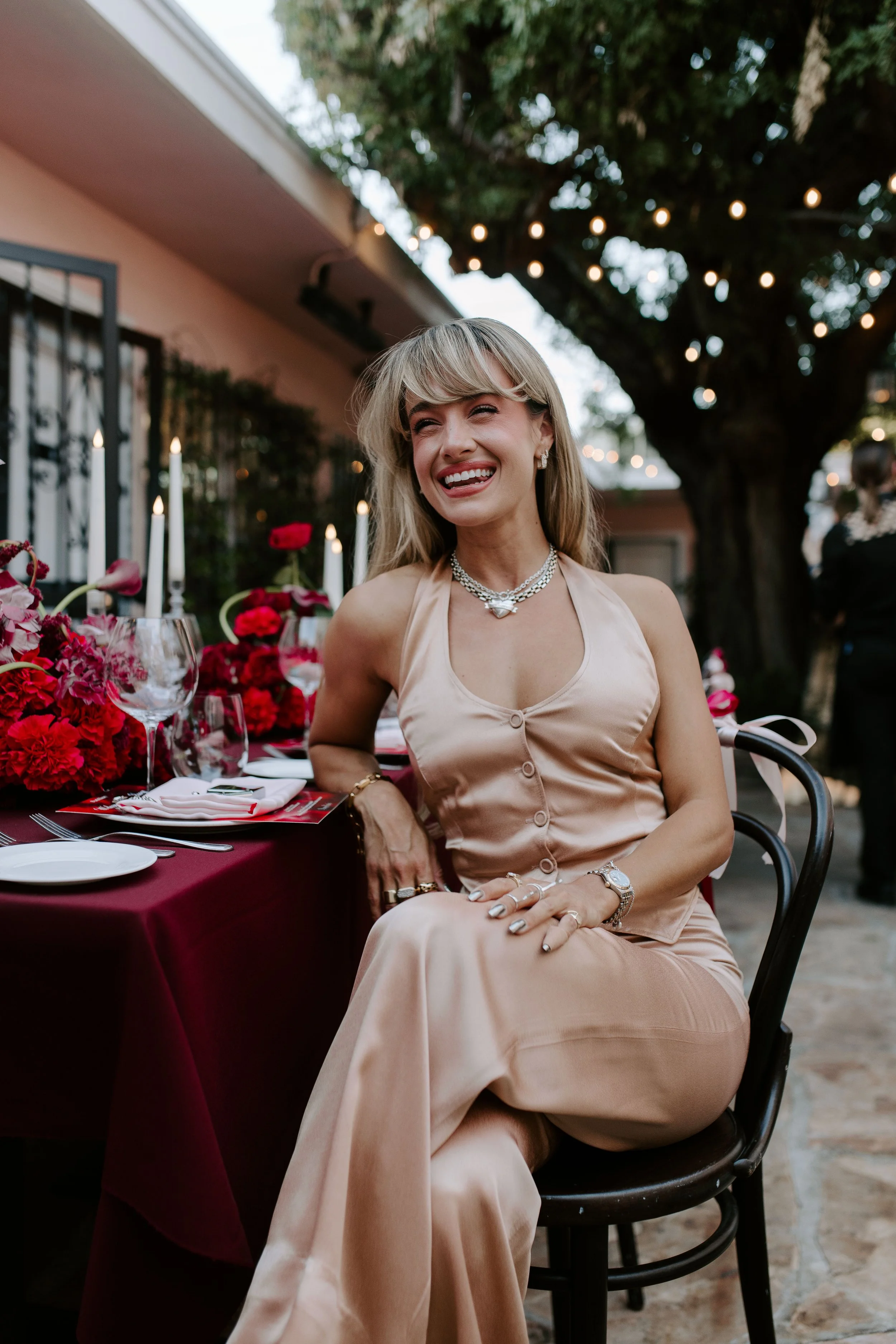 A woman smiling and wearing a beige satin dress, sitting at an outdoor table with red flowers, wine glasses, and candles, under a string of lights and trees.