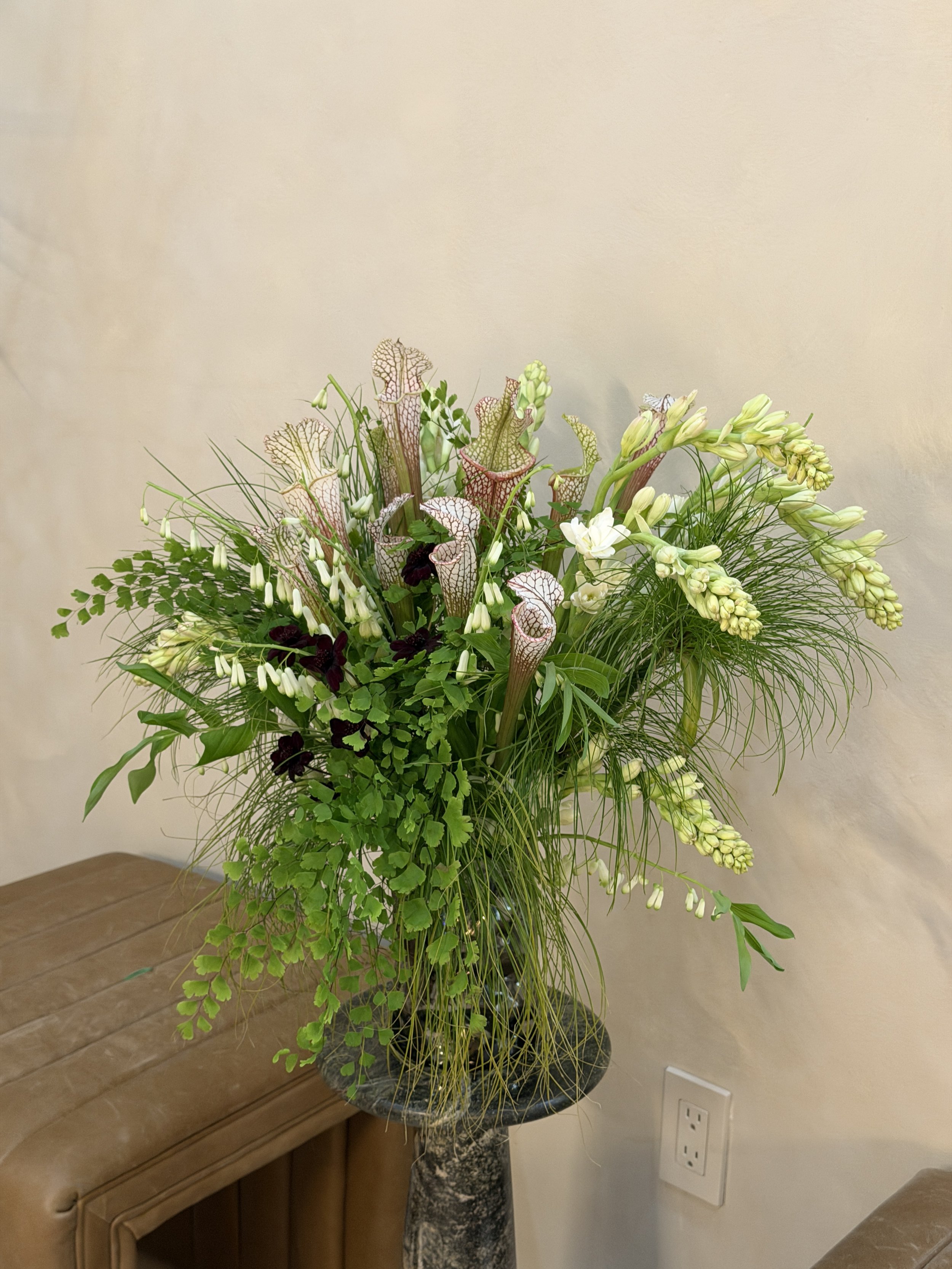 A large, colorful floral arrangement in a black marble vase on a wooden table, featuring tall green foliage and white and burgundy flowers against a beige wall.