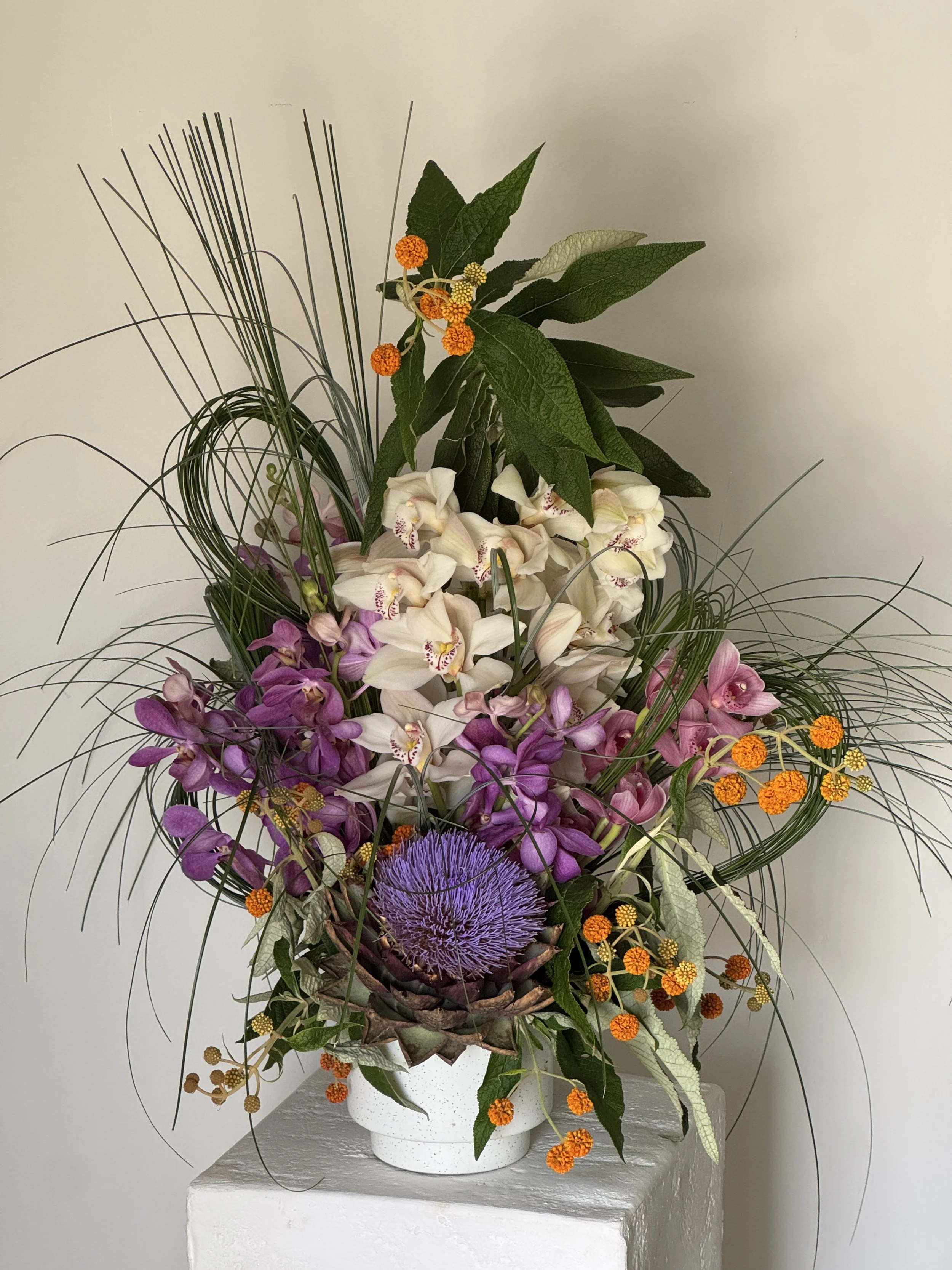 A vibrant floral arrangement in a white vase features purple, white, and pink orchids, with greenery, orange, and yellow accents, and a large purple artichoke flower at the center.