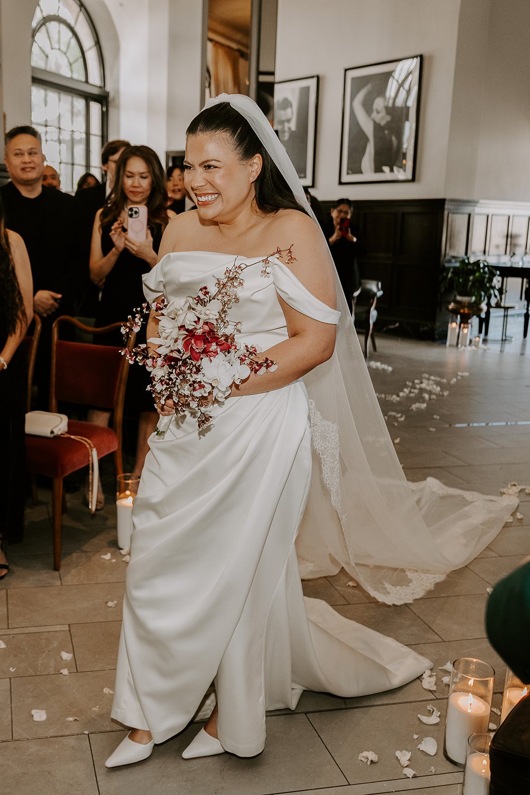 A joyful bride in a white wedding dress holding a floral bouquet, standing in a decorated indoor wedding venue with guests cheering around her.