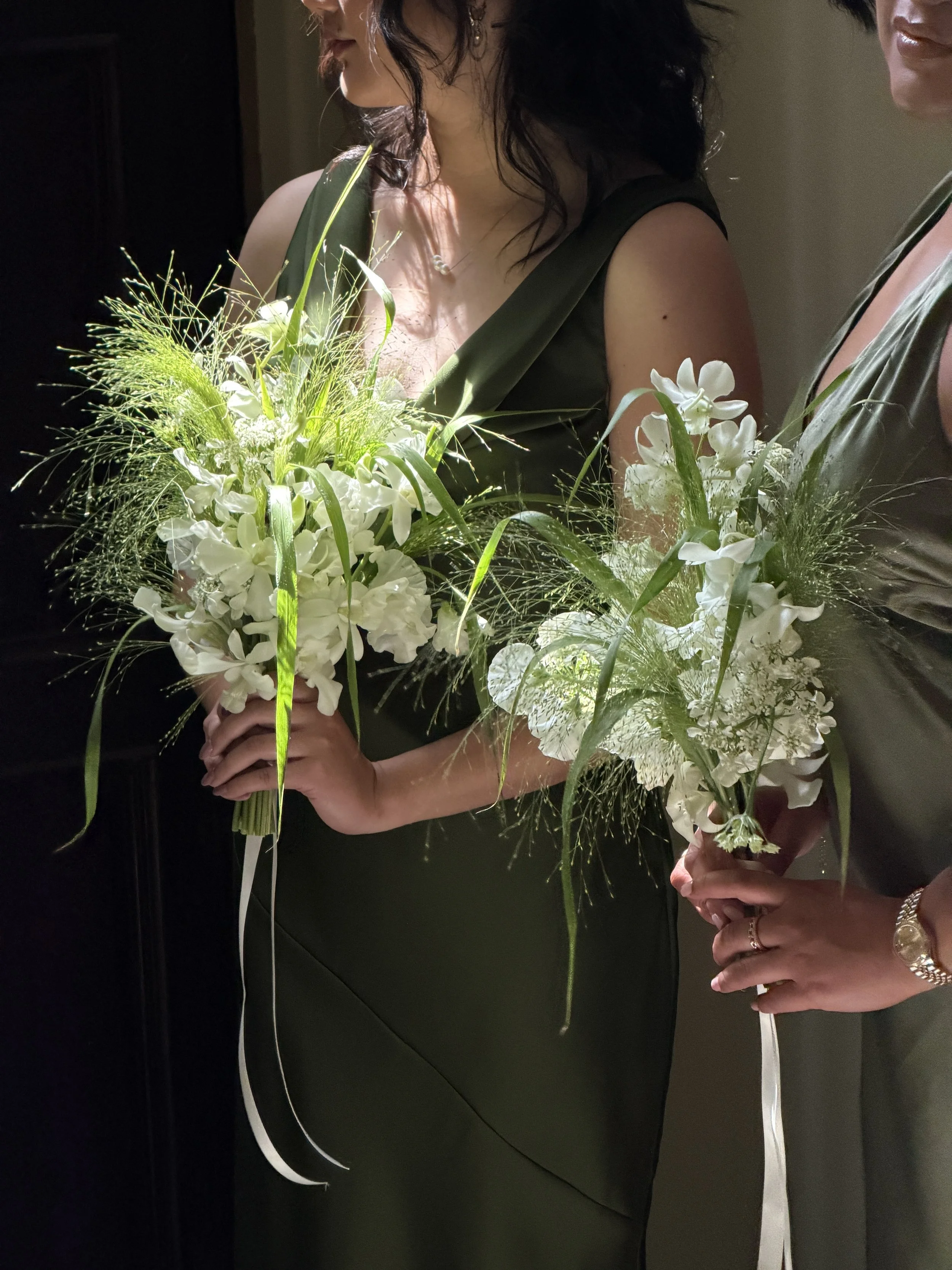 Two bridesmaids in elegant dresses holding bouquets of white flowers with greenery, standing close together in a dimly lit setting.