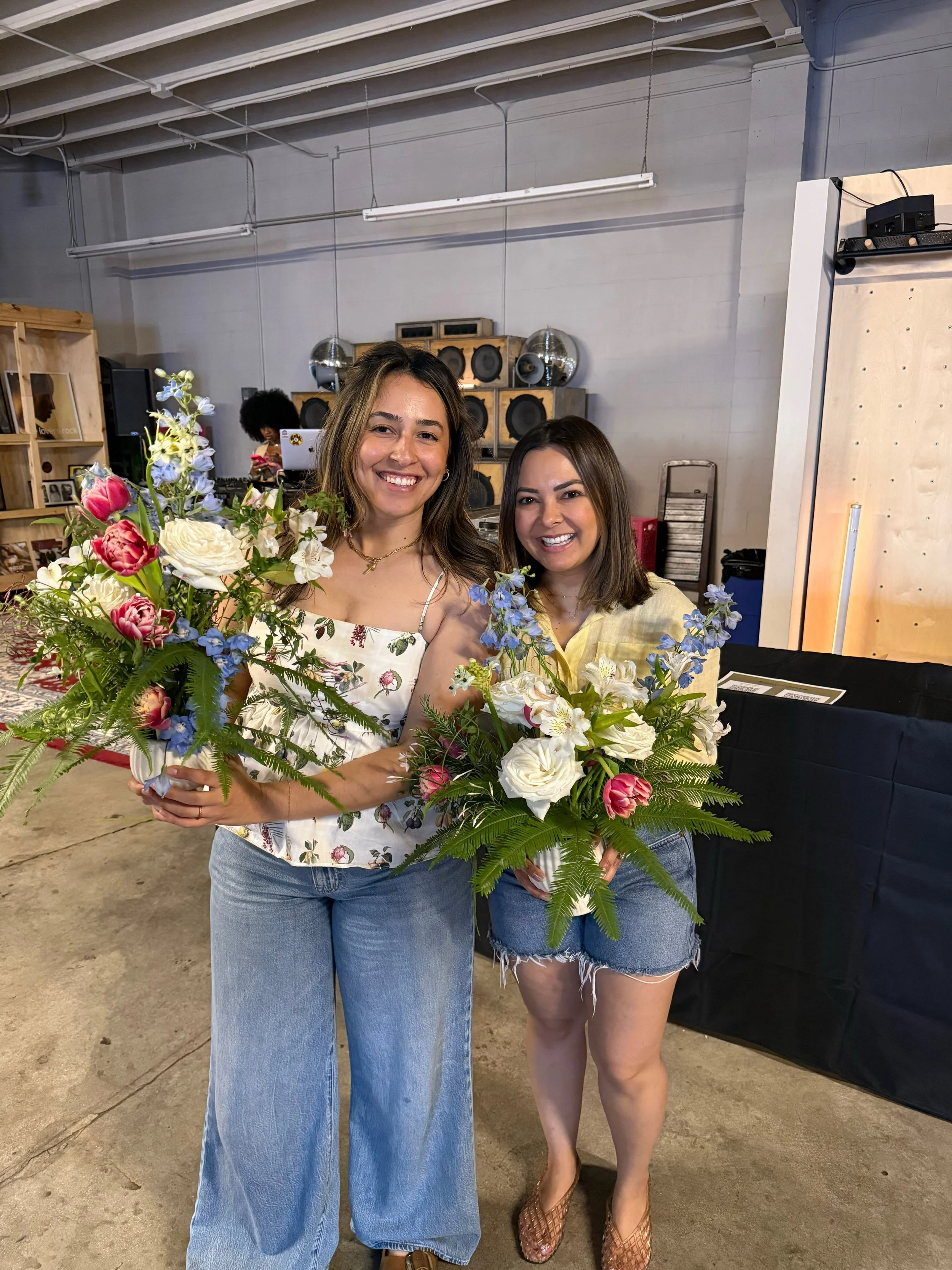 Two women smiling and holding large bouquets of flowers inside a building with a concrete floor, with speakers and a person in the background.