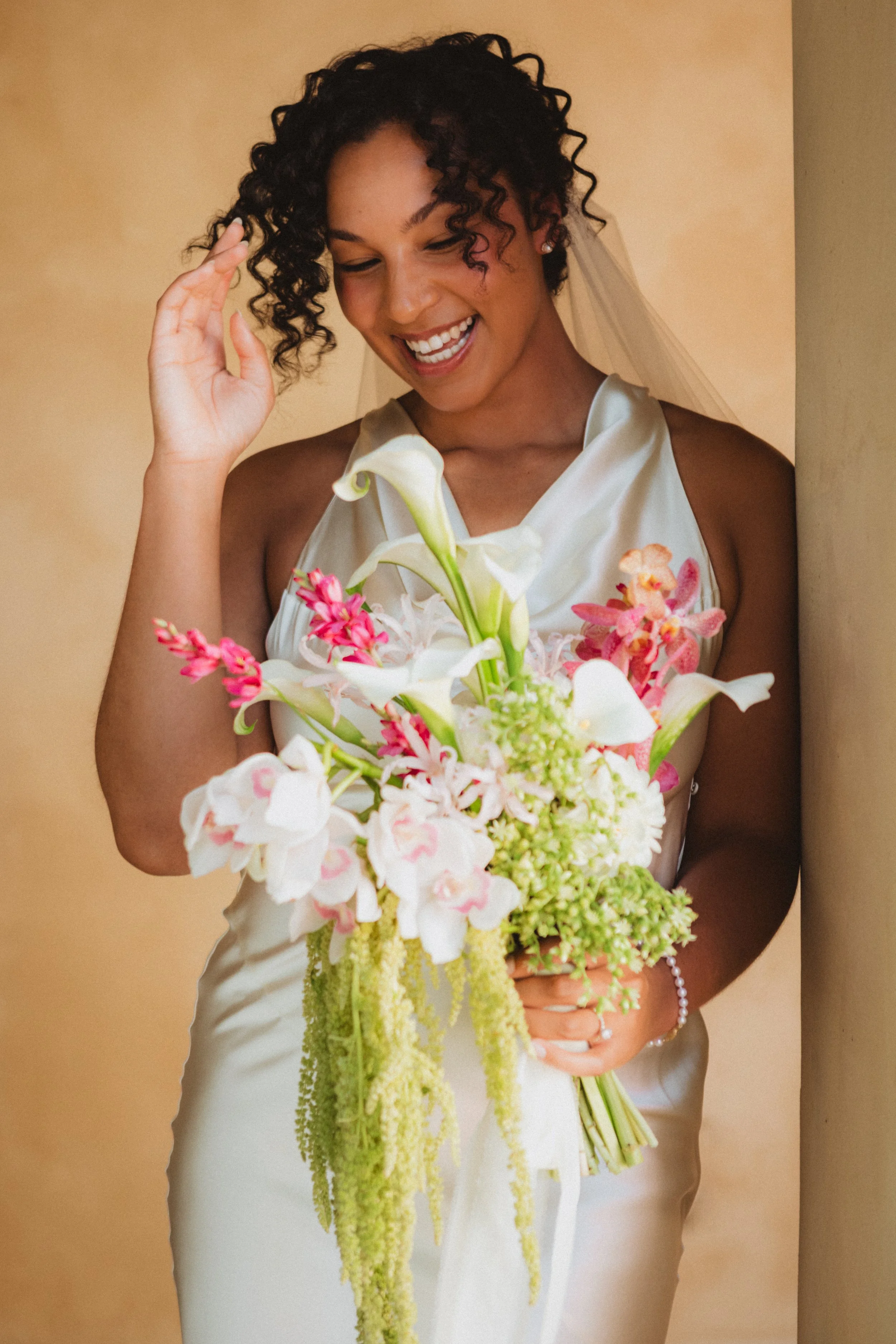 A smiling bride wearing a white dress and pearl earrings, holding a large bouquet of white, pink, and green flowers, standing against a beige wall.