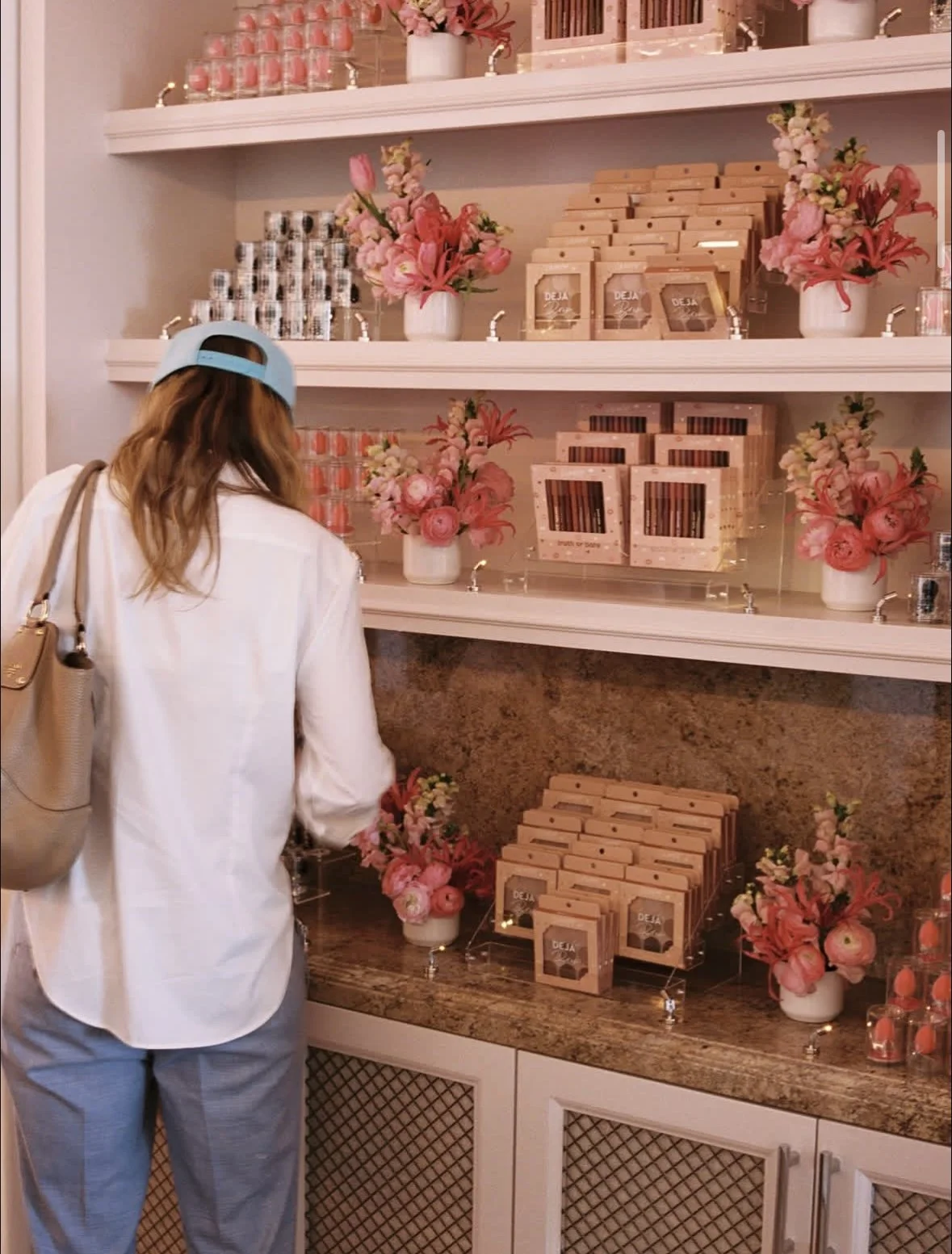 A woman in a white jacket and blue baseball cap shopping in a store decorated with pink flowers and various pink items displayed on white shelves.