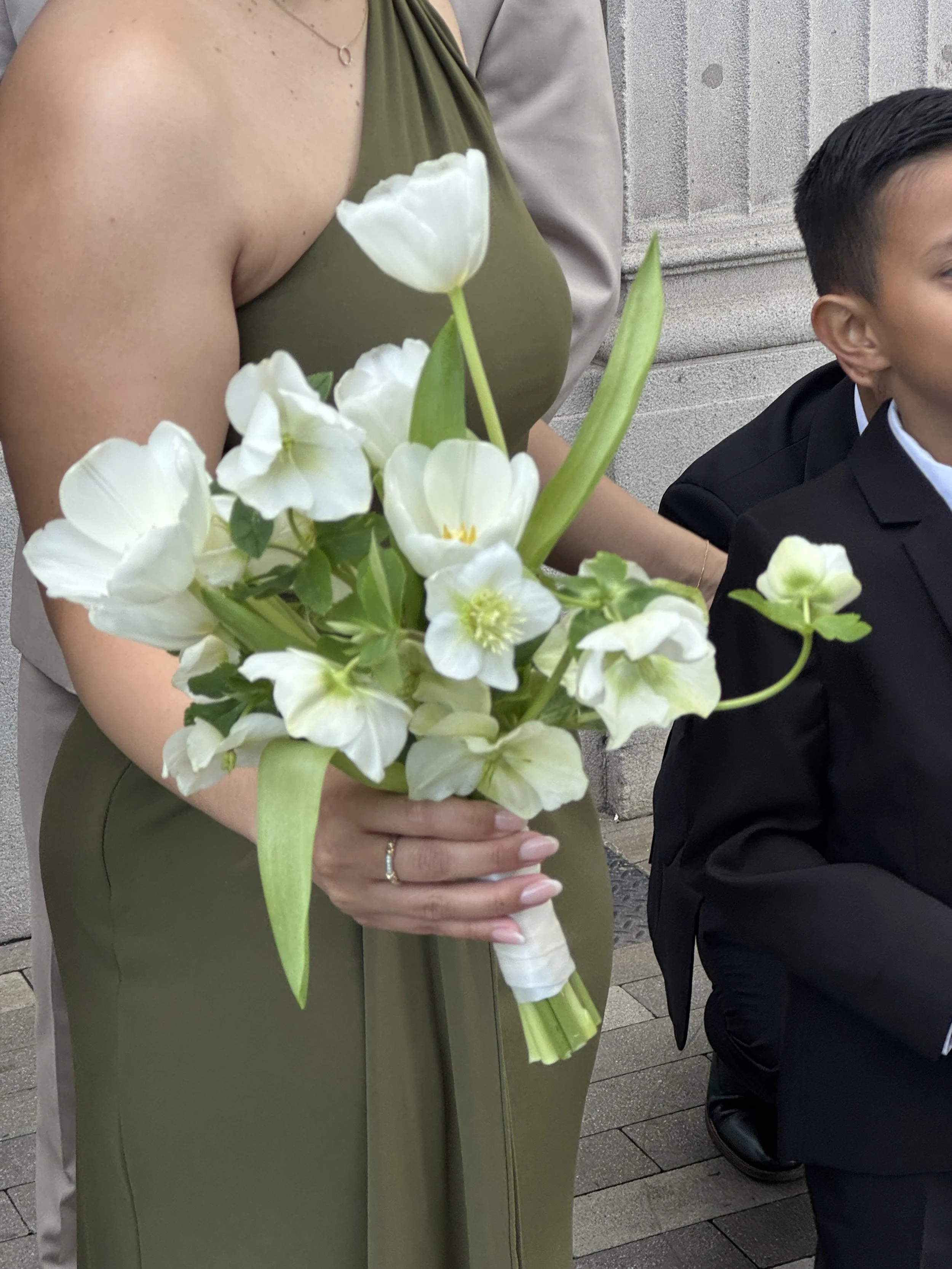 A woman in an olive green dress is holding a bouquet of white flowers, including tulips and other blooms. Next to her, a man in a black suit with short dark hair is partially visible, sitting on a stone step outside.