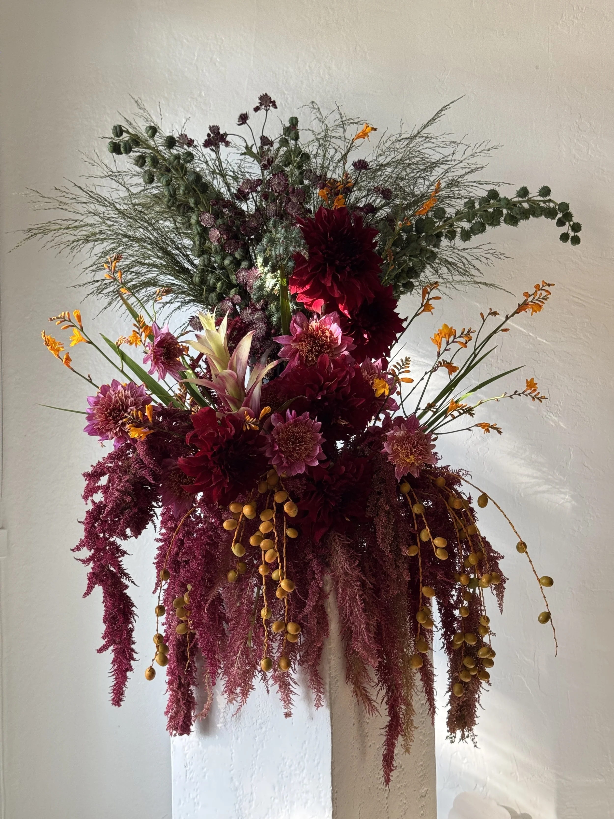 A tall, colorful floral arrangement with burgundy, pink, orange, and green flowers and foliage, hanging vertically against a white wall.