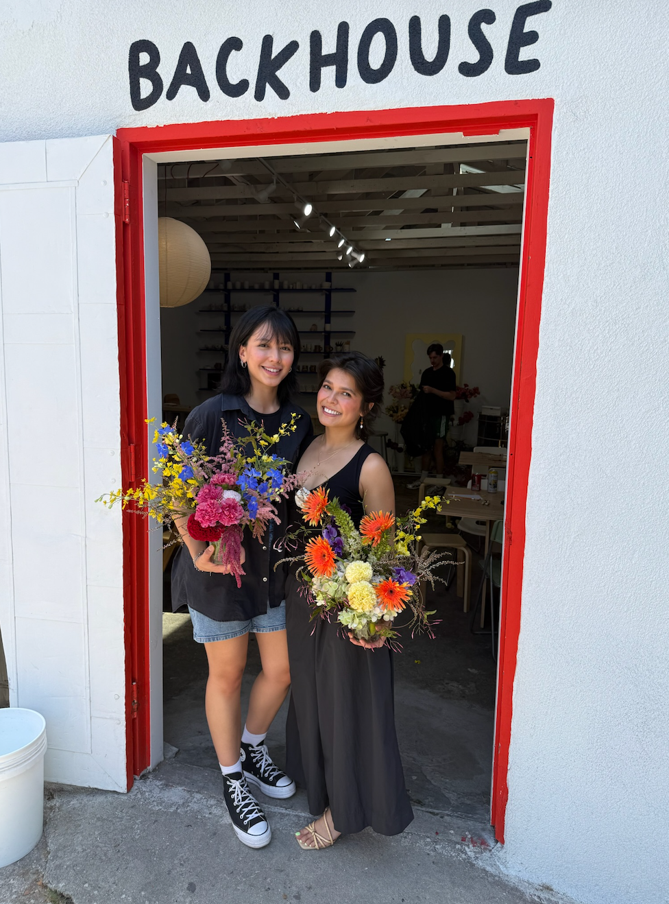Two women standing in front of a backhouse with a sign above the door, holding colorful bouquets of flowers, smiling at the camera.