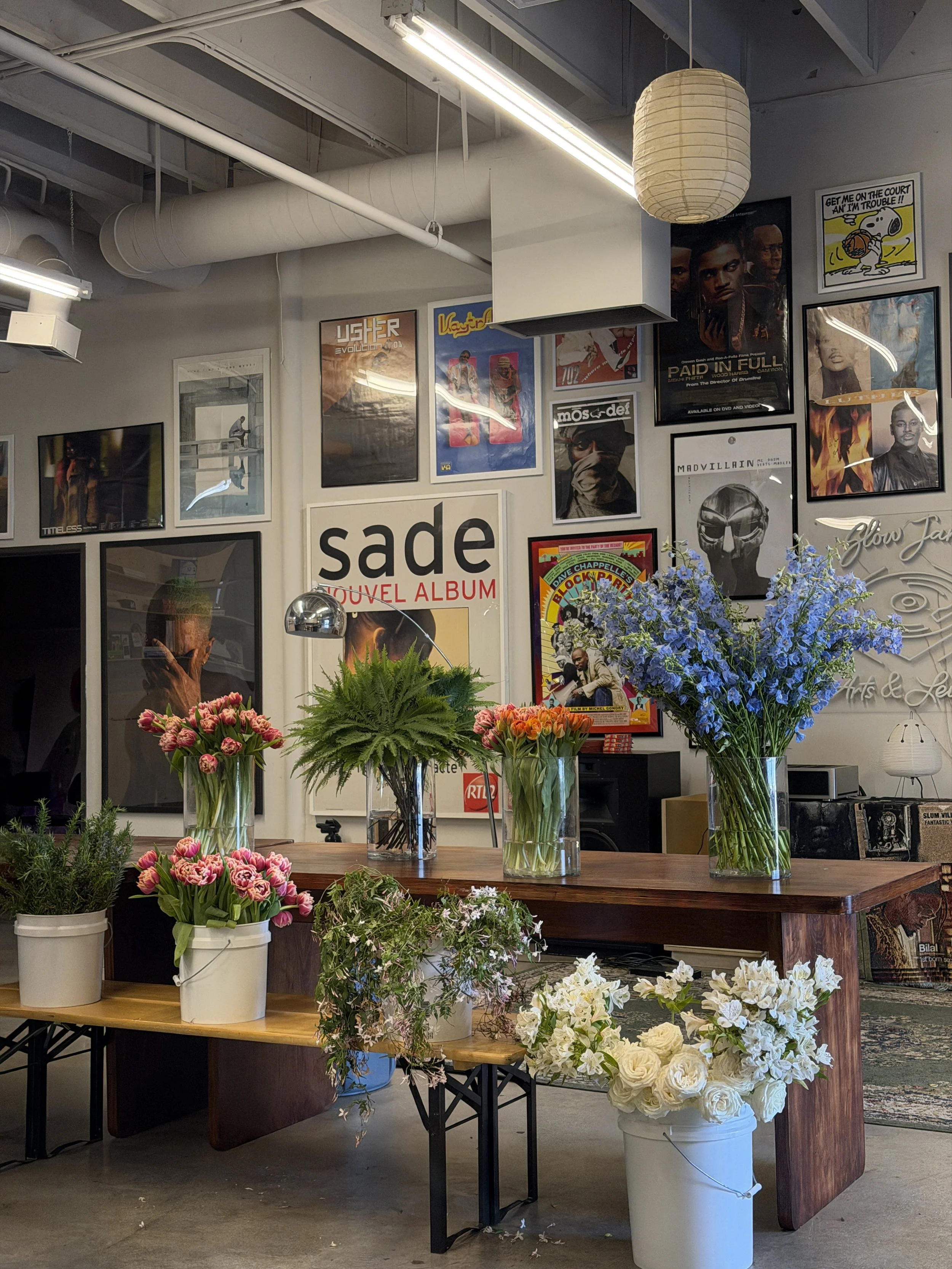 Several glass vases filled with flowers, including pink tulips, white roses, purple delphiniums, and assorted greenery, arranged on a wooden table inside a showroom or floral shop. The background features framed posters and artwork on the wall.