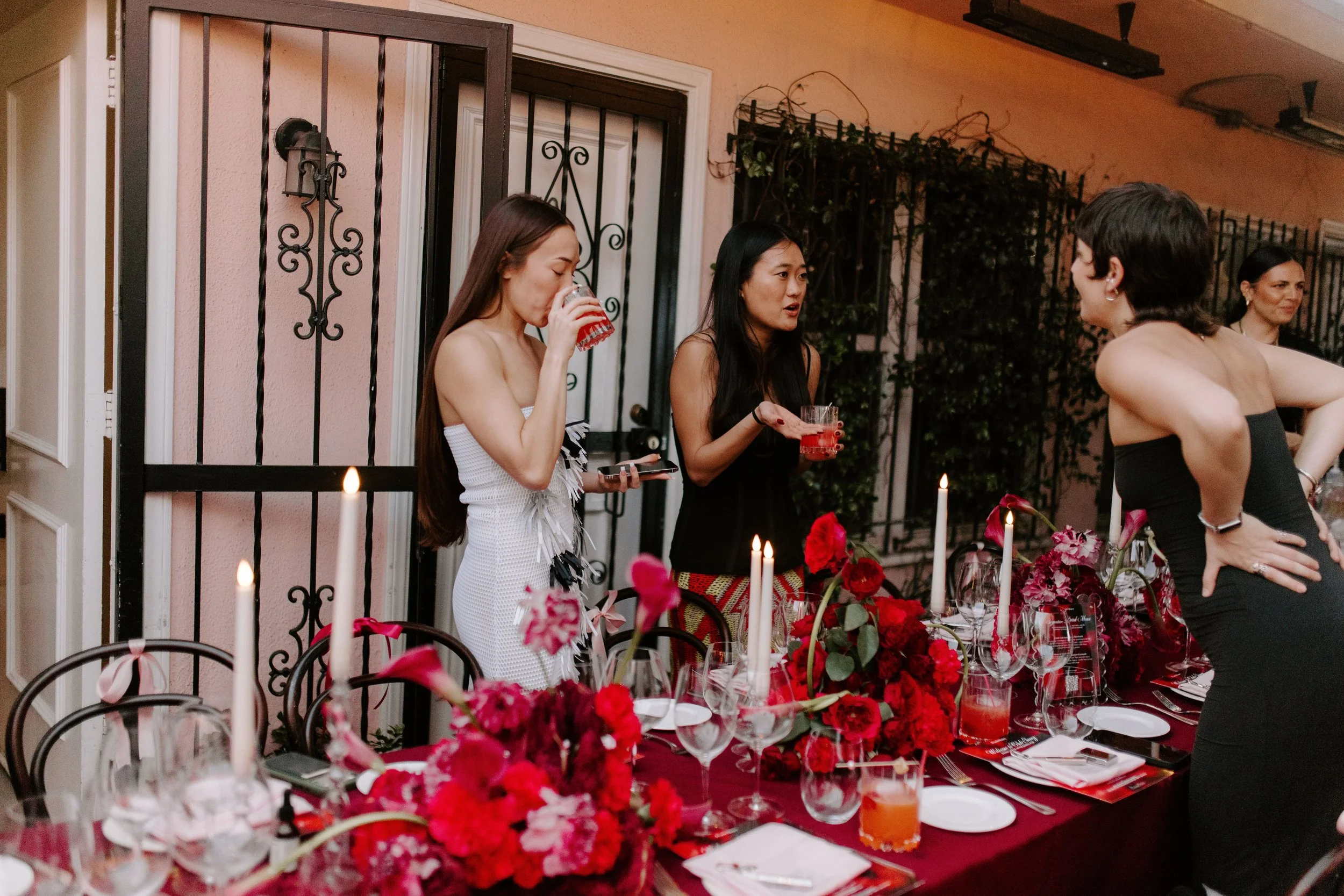 A group of women at a dinner party, standing around a table decorated with red flowers, candles, and glassware, inside a room with pink walls, black iron gate, and greenery outside.