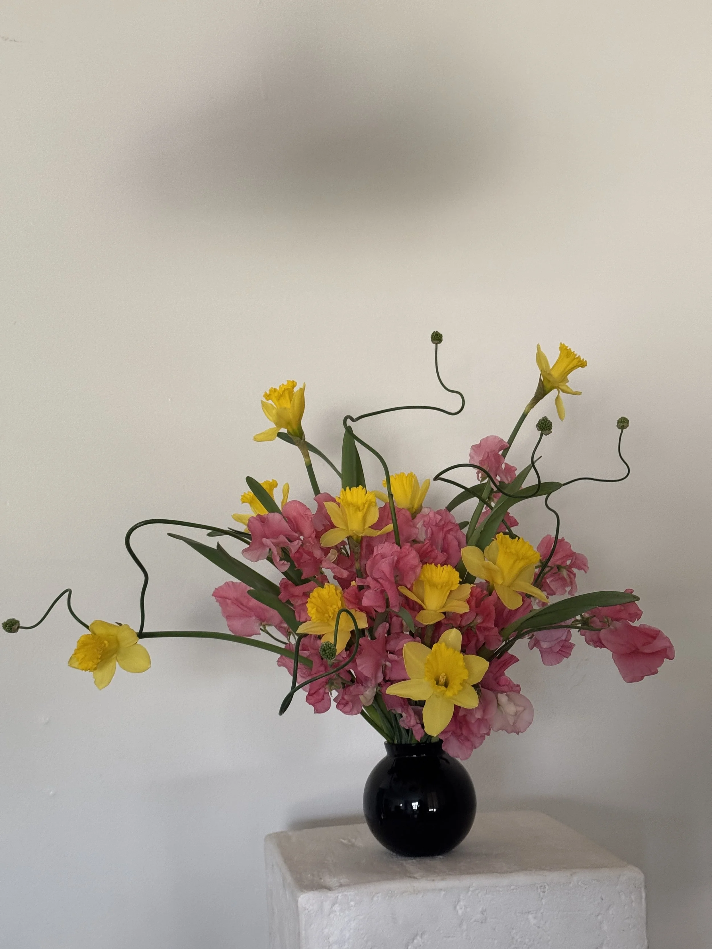 A bouquet of pink and yellow flowers in a black vase on a white pedestal against a plain white wall.