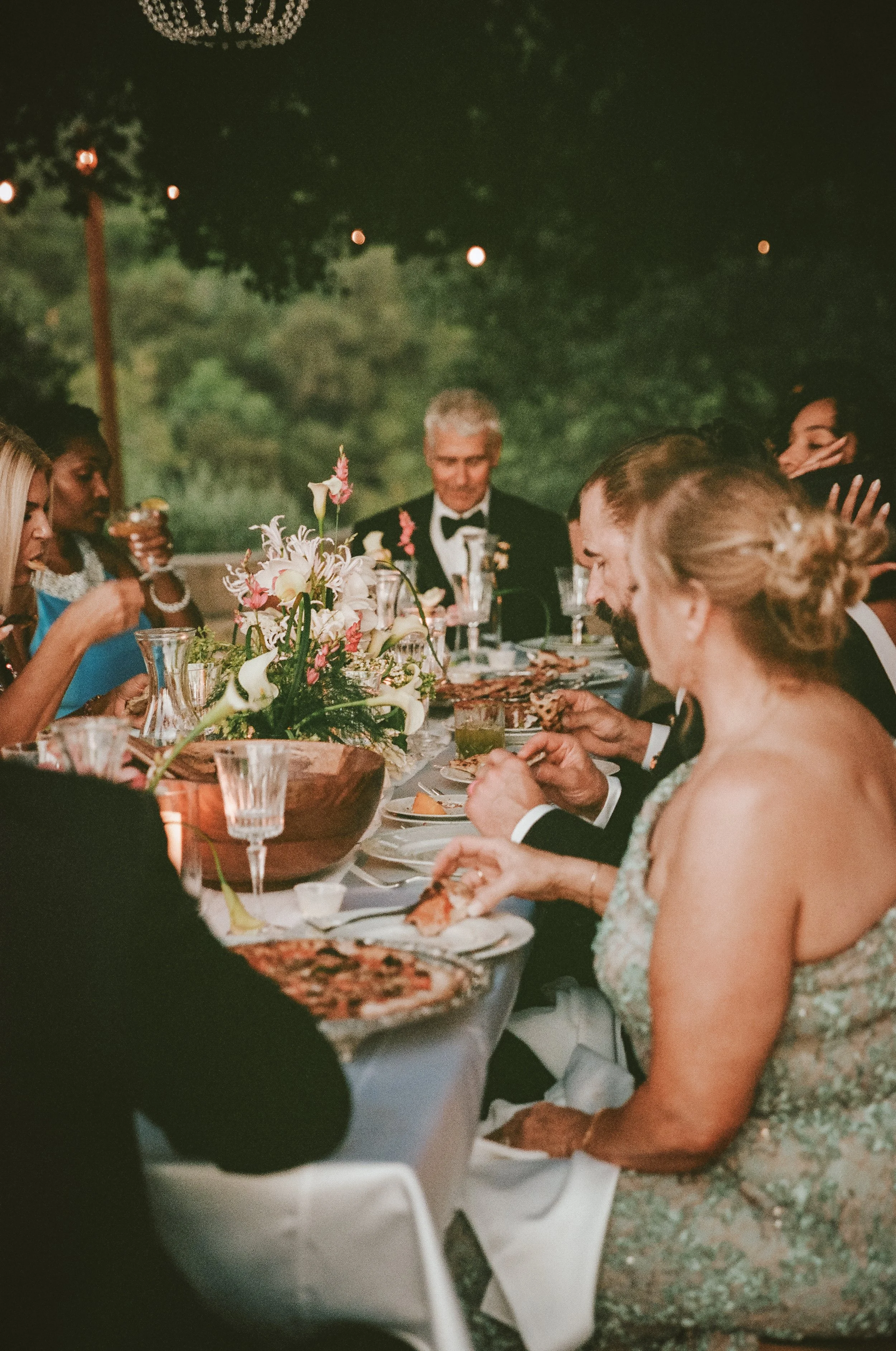 A group of people dressed in formal attire are sitting around a long table at an outdoor dinner party at night. There are floral centerpieces and glassware on the table, and one person is holding a glass. The background features trees and string lights.