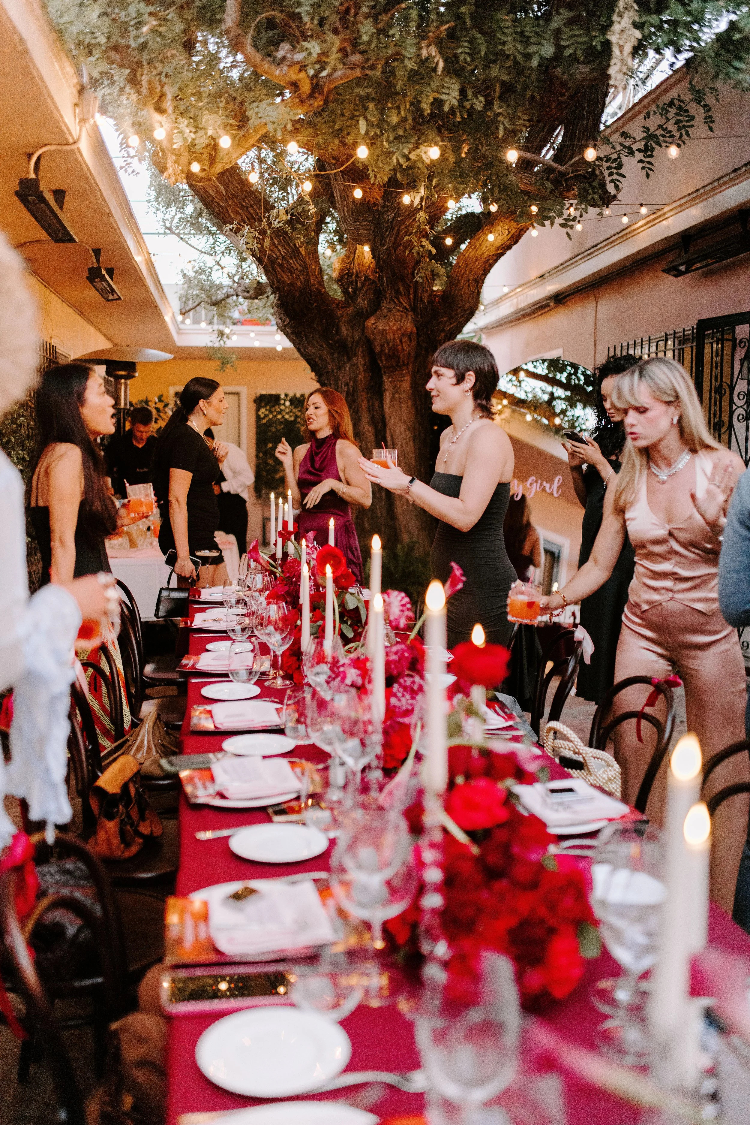 A group of people gathered at a festive dinner party outdoors under a large tree decorated with string lights. The table is set with red tablecloths, floral centerpieces, candles, plates, glasses, and utensils. The guests are standing, chatting, and holding drinks, dressed in formal or semi-formal attire.