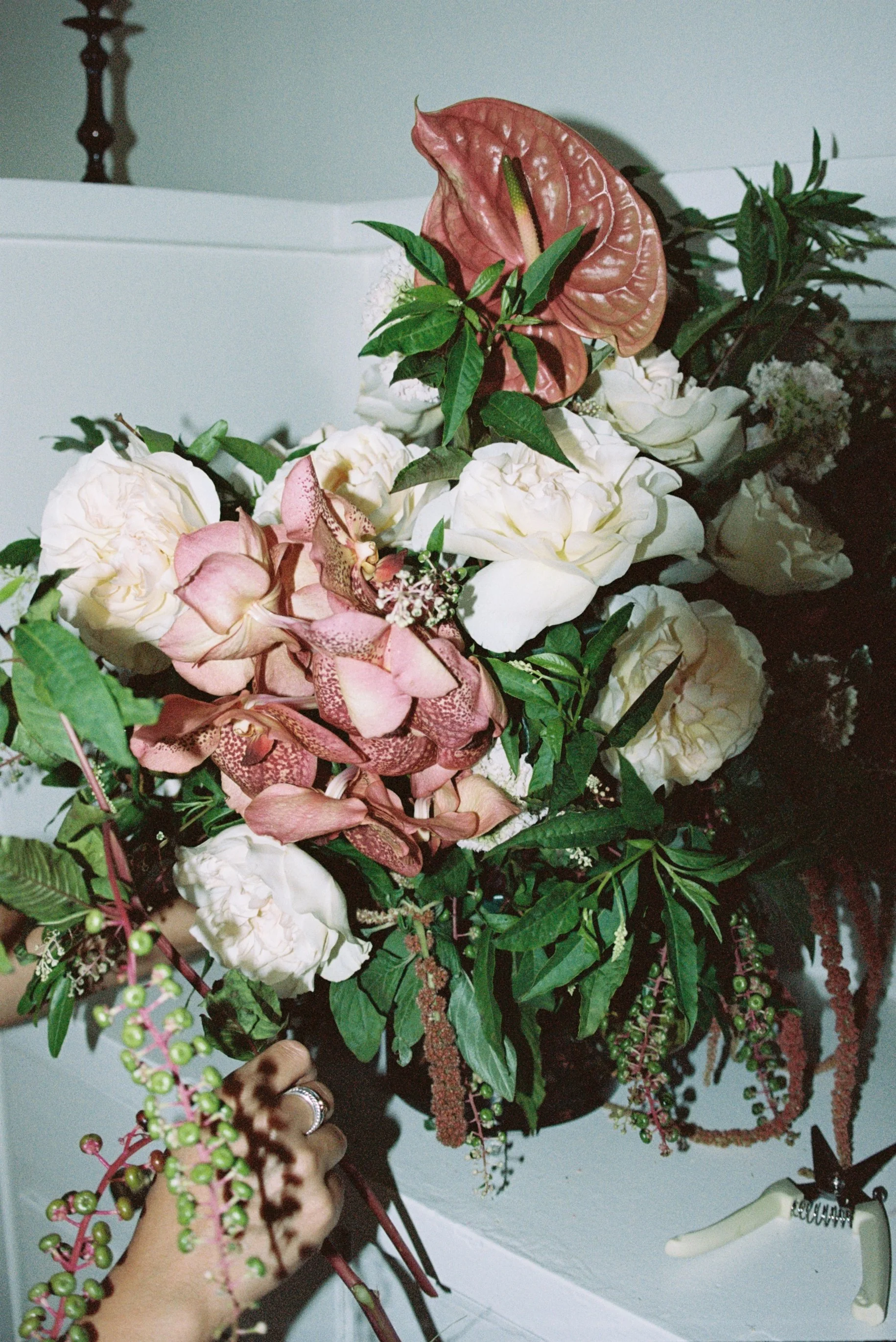A large bouquet of various flowers including white roses, pink orchids, and a pink anthurium, with green leaves and berries, with a hand holding the bouquet.