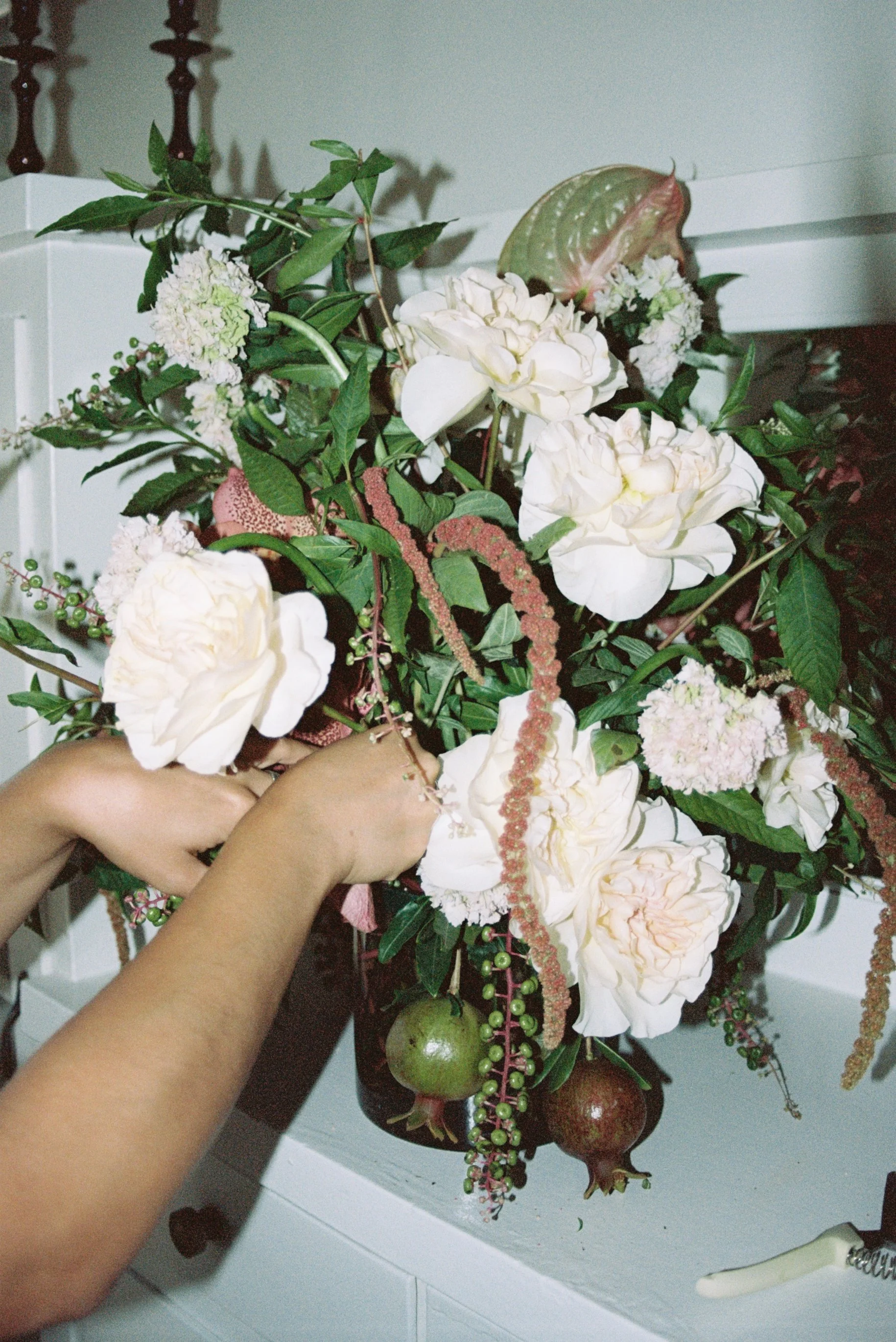 A woman arranging a bouquet of flowers with white blooms, green leaves, and hanging pink and green pomegranate fruits.