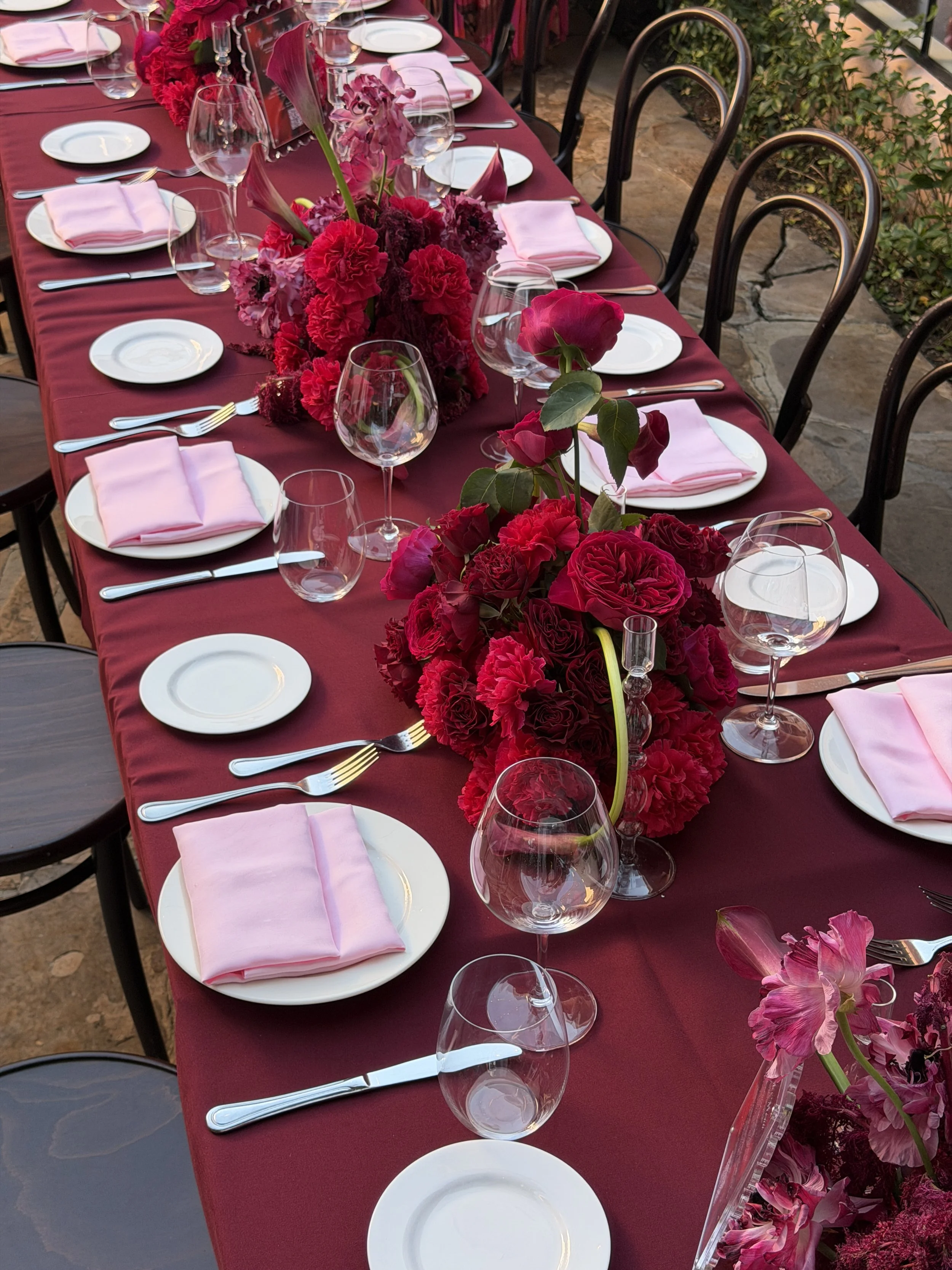 A long dining table decorated with pink and red flowers, set with white plates, pink napkins, silverware, and empty wine glasses. The table has a maroon tablecloth, and the chairs are black. The setting appears elegant and prepared for a special event or celebration.