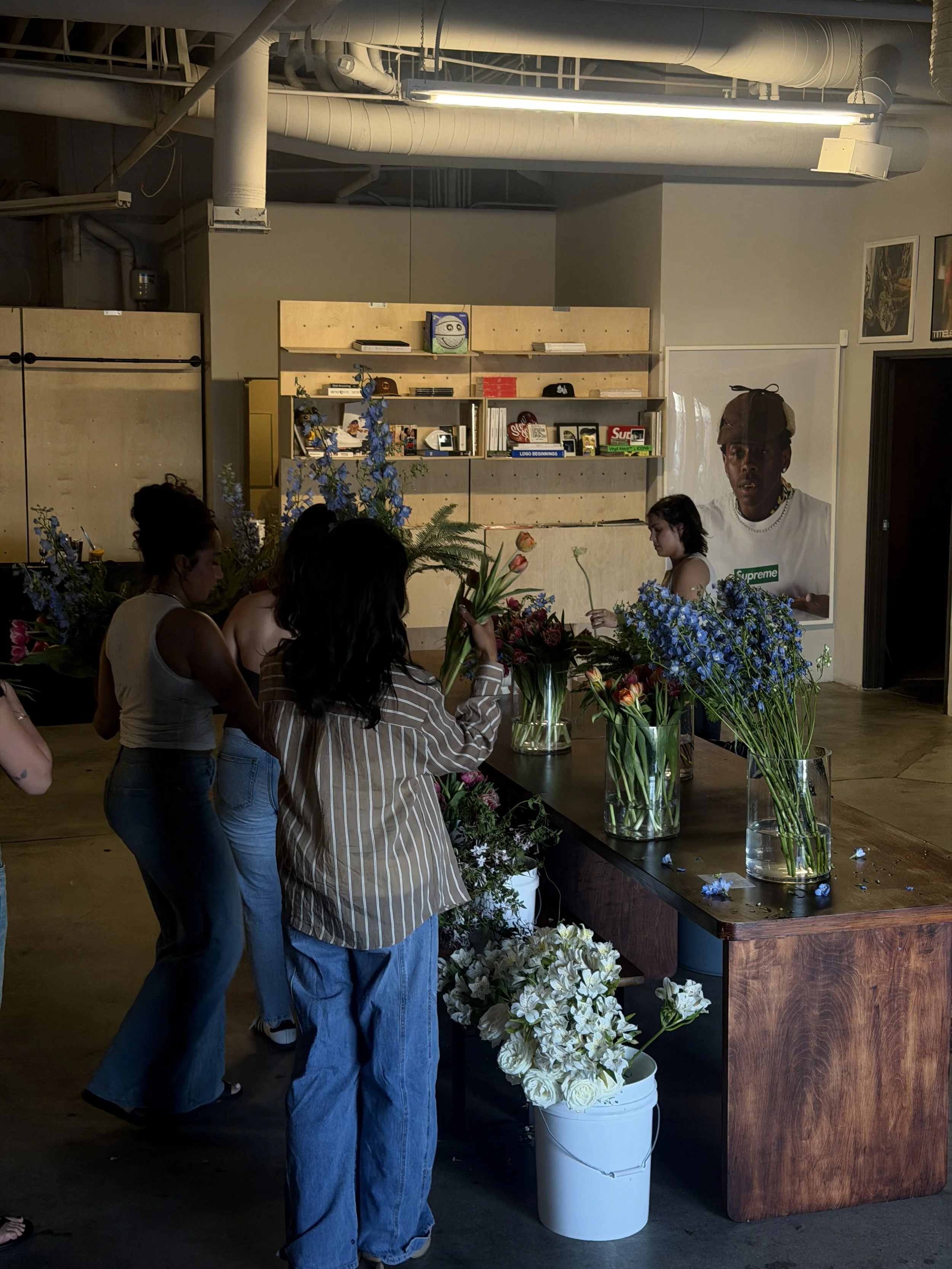 People gathering around a table arranging various flowers in vases inside a room with a shelf filled with books and artwork on the wall.
