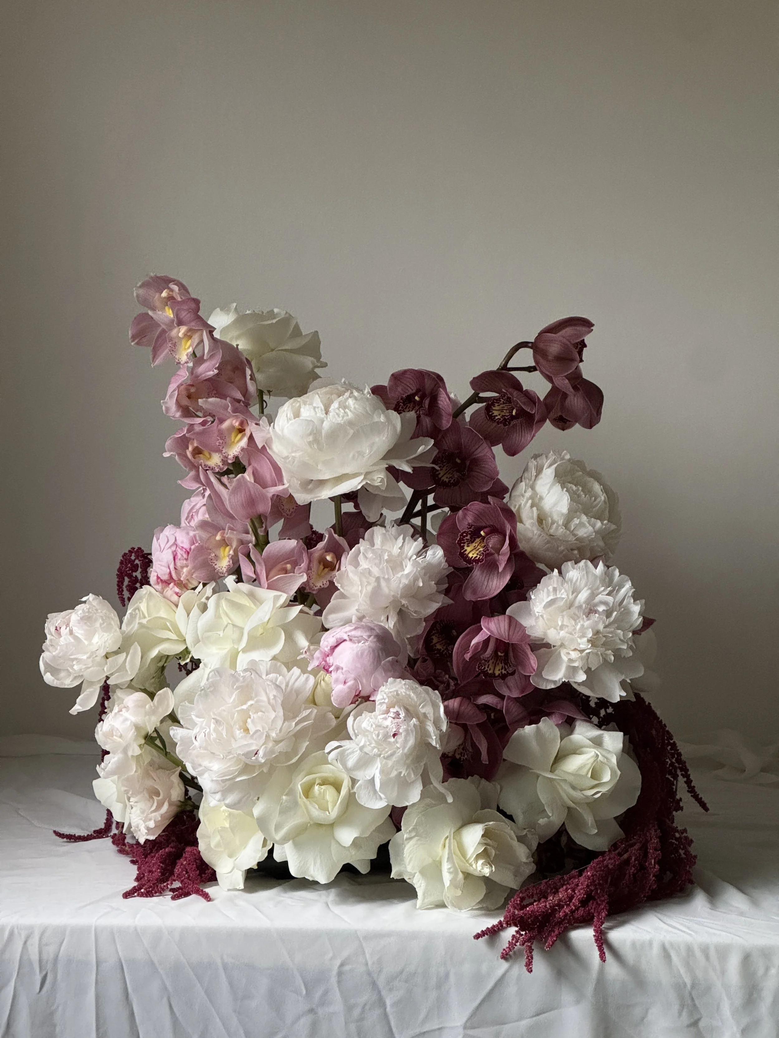 A floral arrangement of white peonies, pink orchids, and purple flowers on a white table with a plain gray background.
