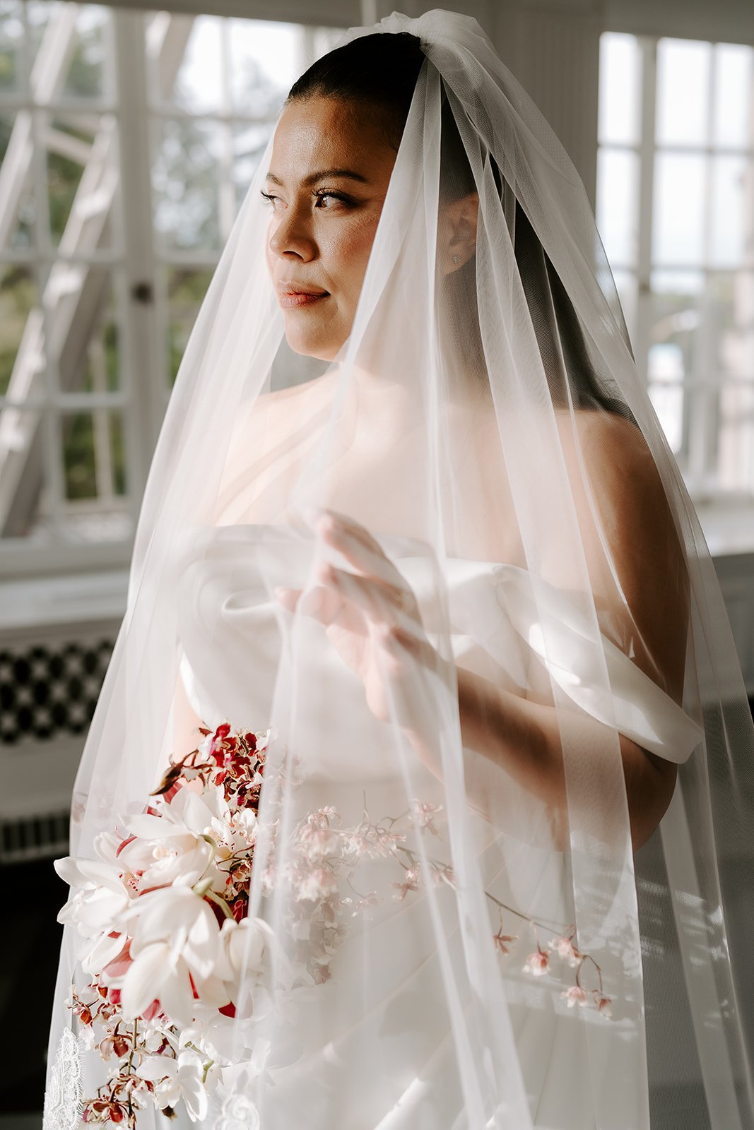 A bride with dark hair styled in an updo, wearing a white strapless wedding gown, a veil, and holding a bouquet of white and red flowers, standing indoors near large windows.