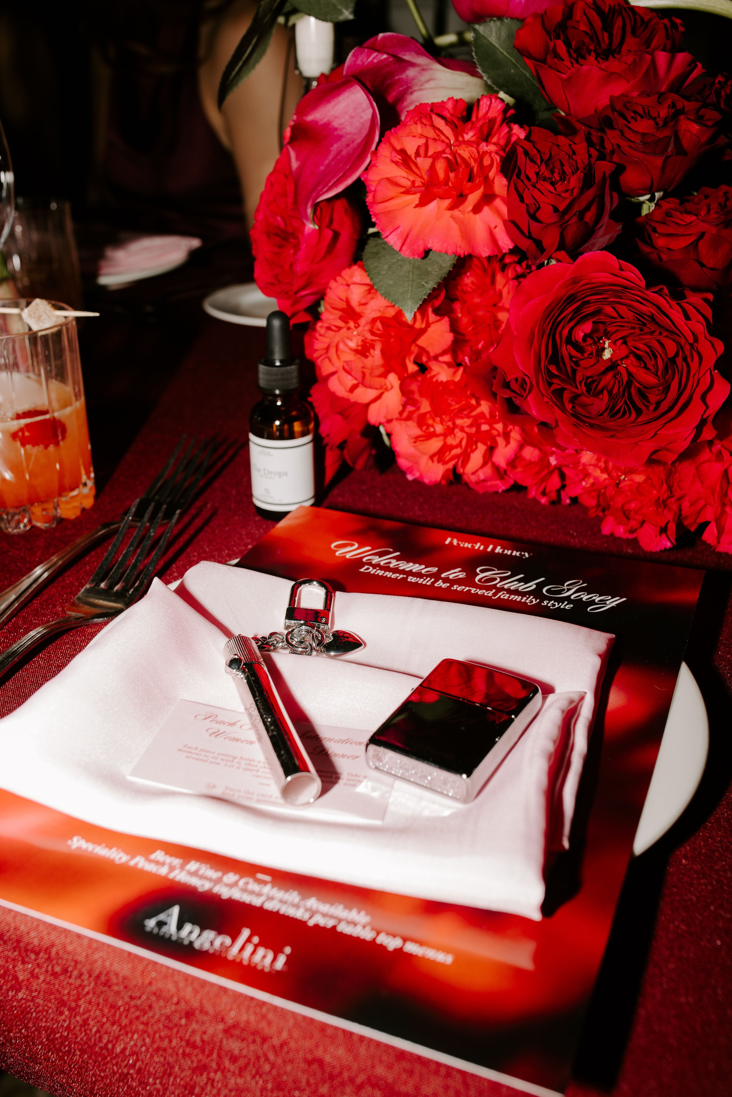Close-up of a table setting with a stack of red flowers including roses and carnations, a white napkin with a black pen, a small glass container, a black box, a menu or program, and a bottle of essential oil, all arranged on a dark red tablecloth.