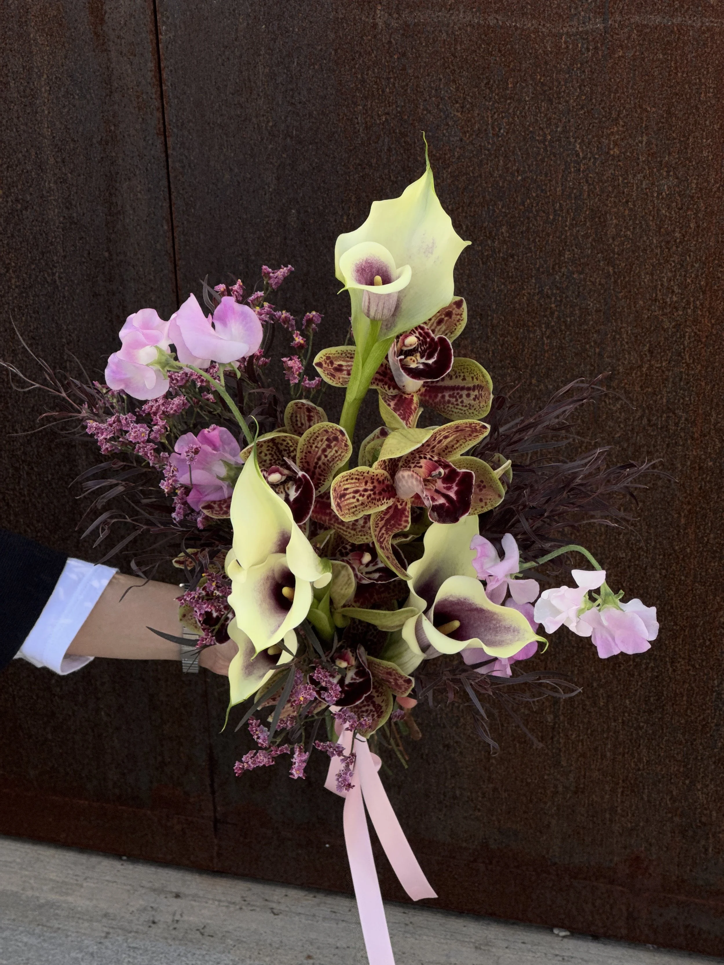 A floral arrangement with pink, white, and purple flowers, including calla lilies and orchids, with a pink ribbon at the bottom, placed against a dark brown background.