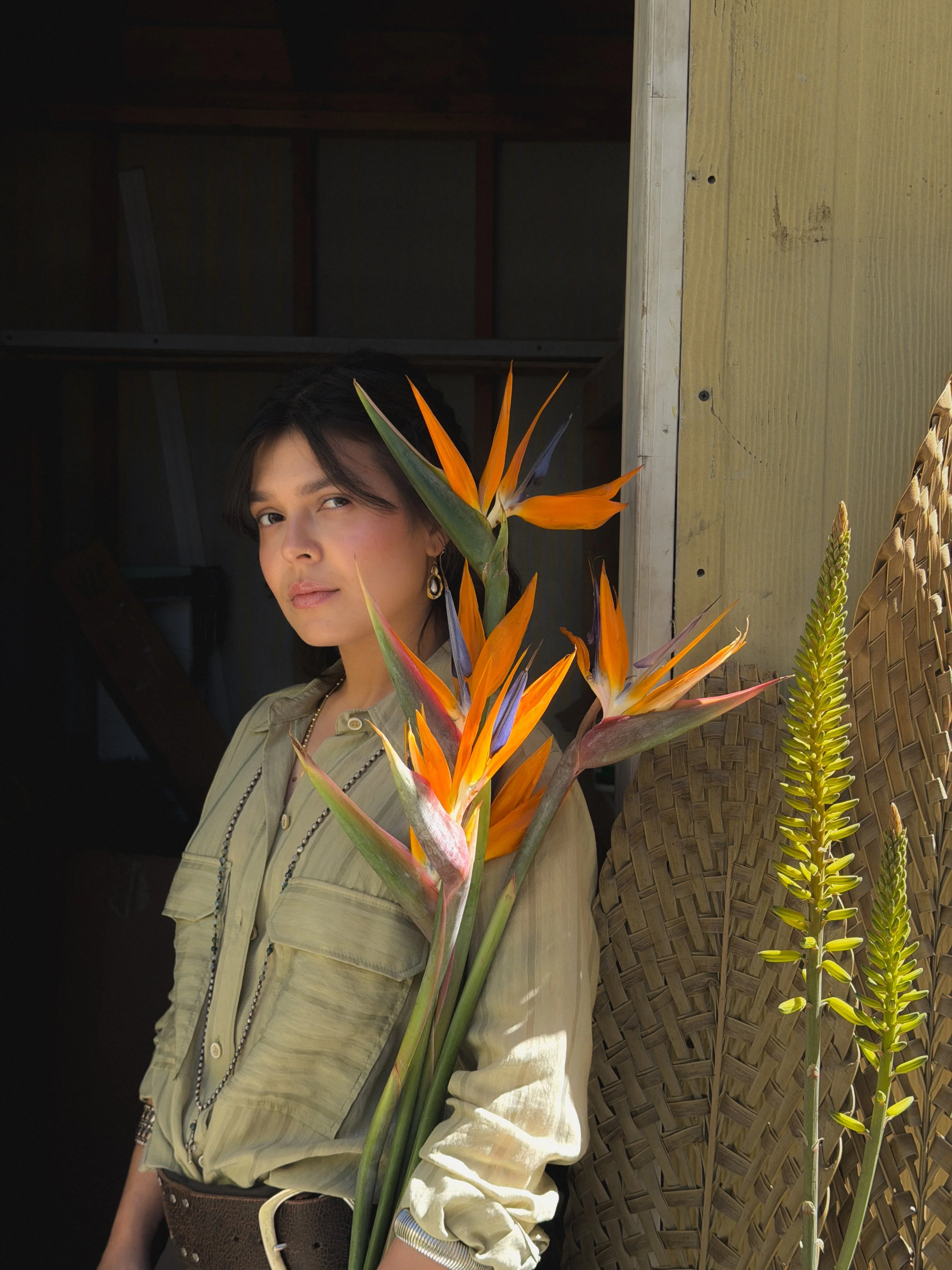 A young woman with short dark hair, wearing a light-colored button-up shirt, stands beside a woven basket and tropical flowers, in a partially open wooden shed.