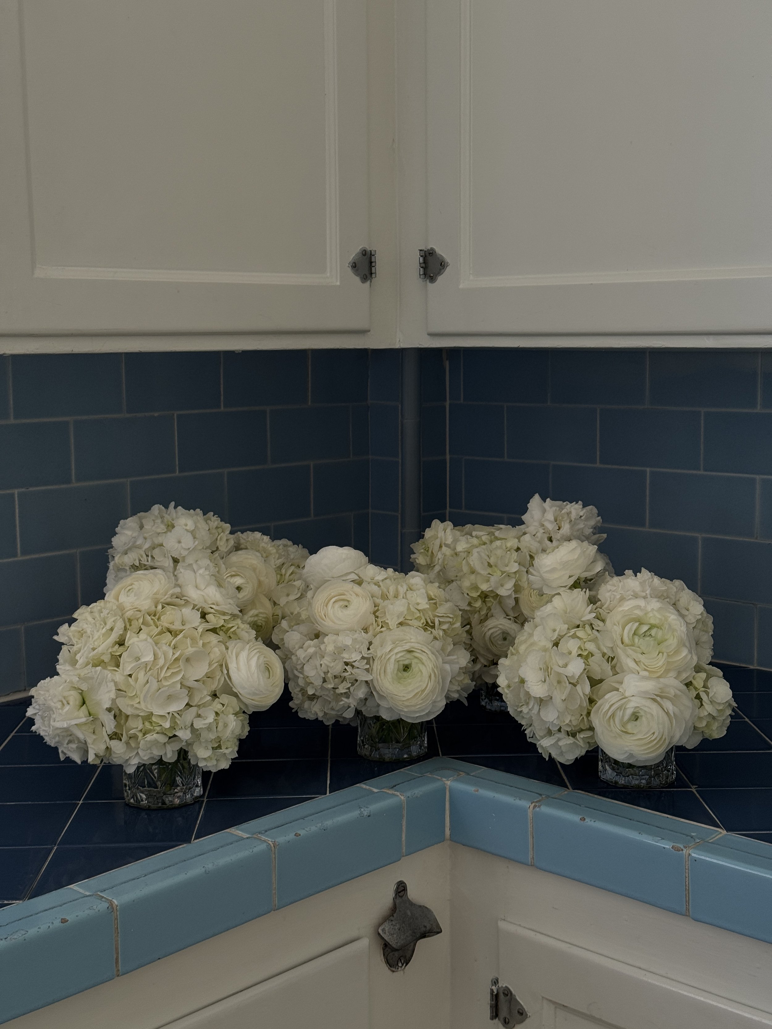White floral arrangements in glass vases placed on blue tiled surface below white kitchen cabinets.