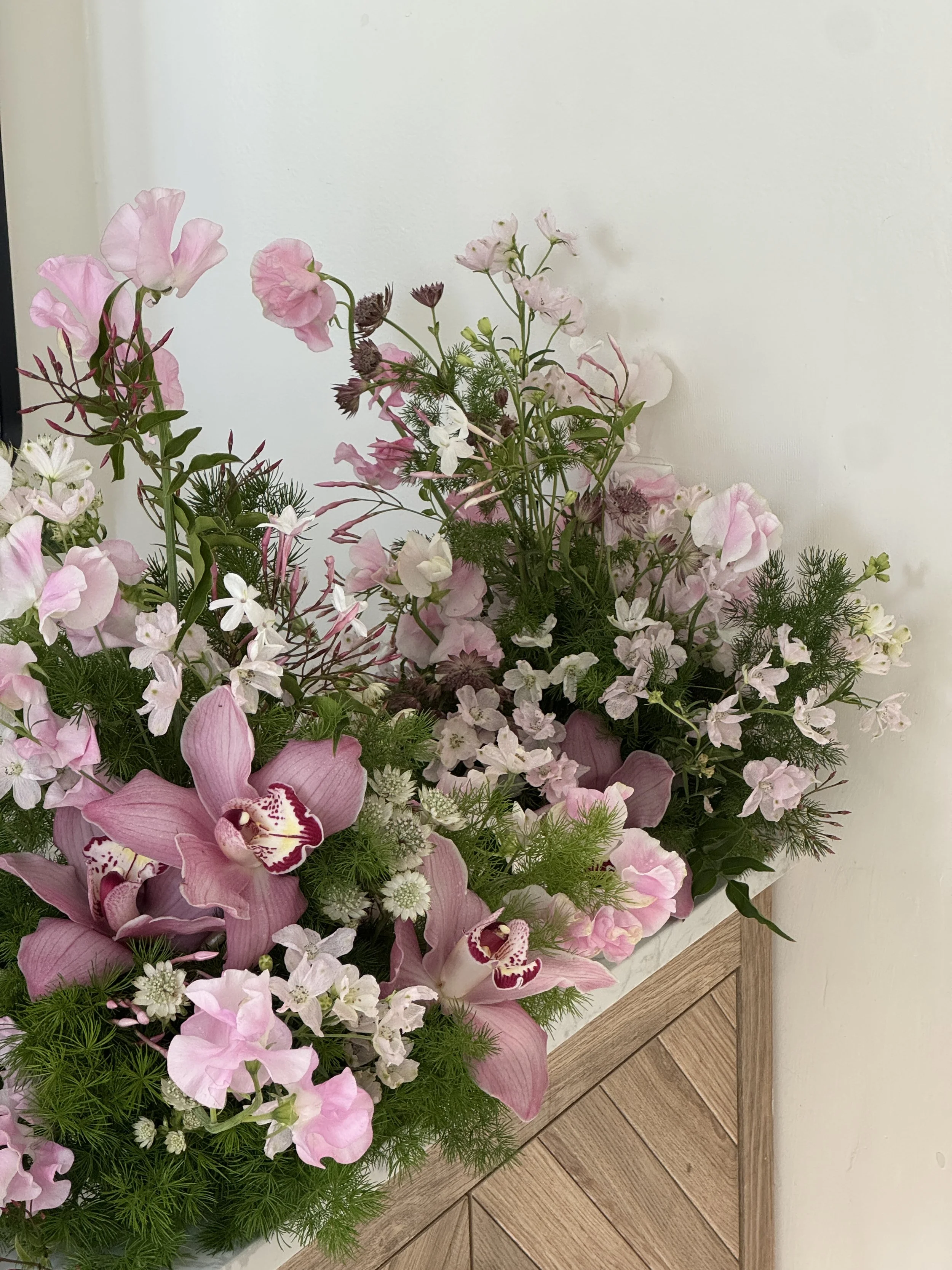 A floral arrangement with pink, white, and purple flowers placed on a wooden stand against a plain wall.