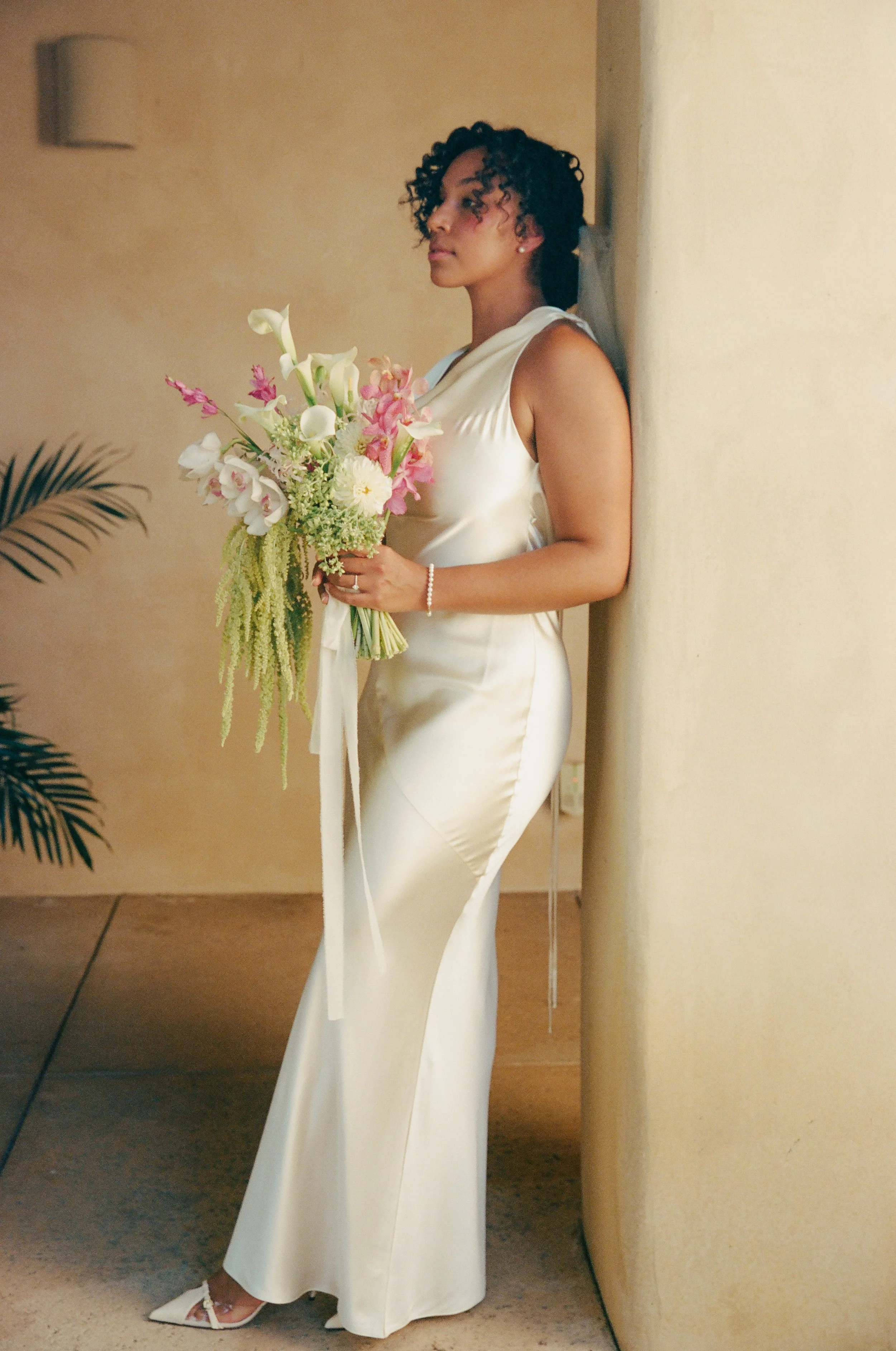 Woman in a white dress holding a bouquet of flowers while leaning against a yellow wall.