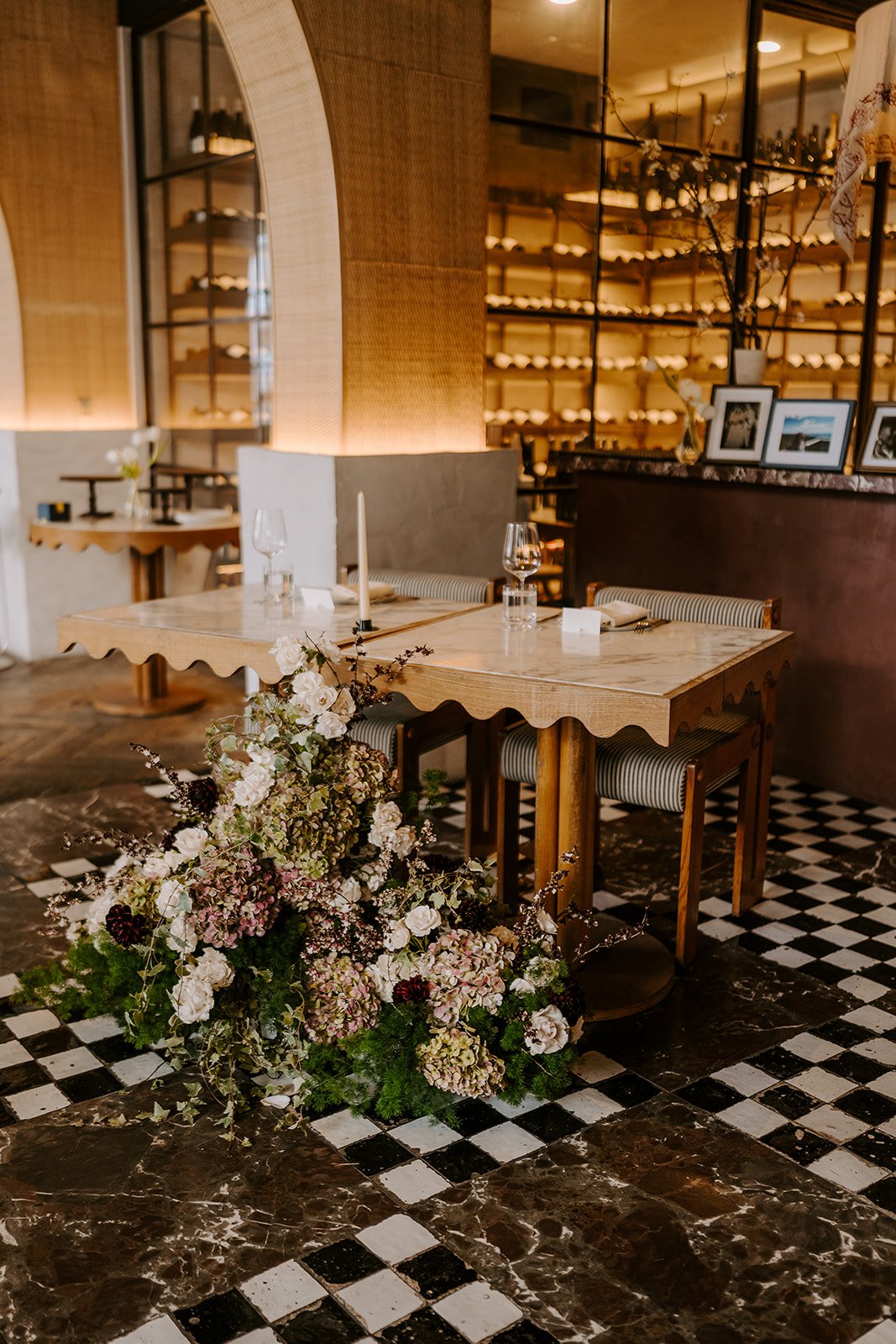 A restaurant interior with wooden tables, floral arrangements, wine glasses, framed pictures, and a shelf with bottles in the background.