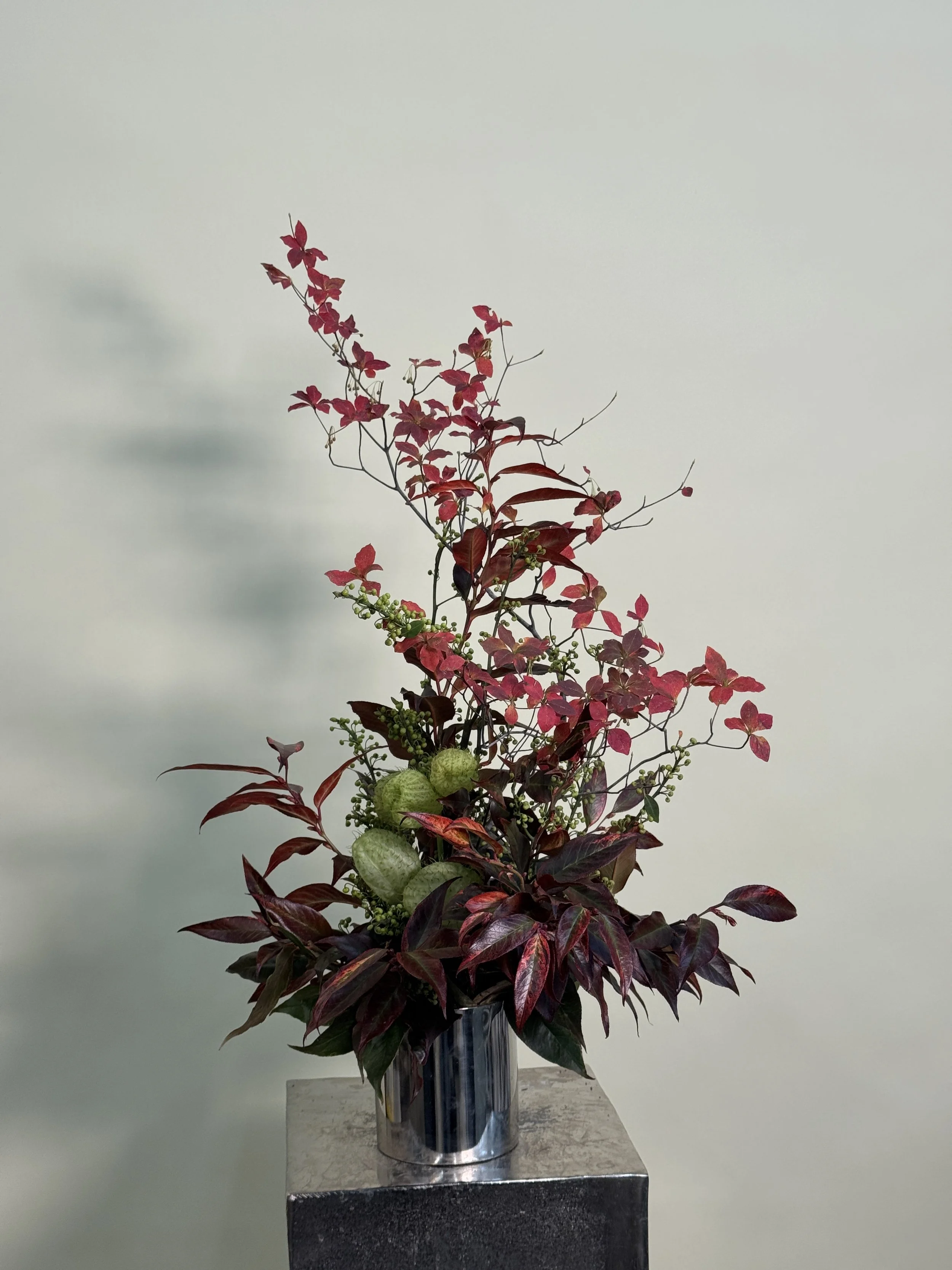 A floral arrangement with red leaves, green seed pods, and dark purple foliage in a shiny metallic vase on a marble pedestal against a plain wall.
