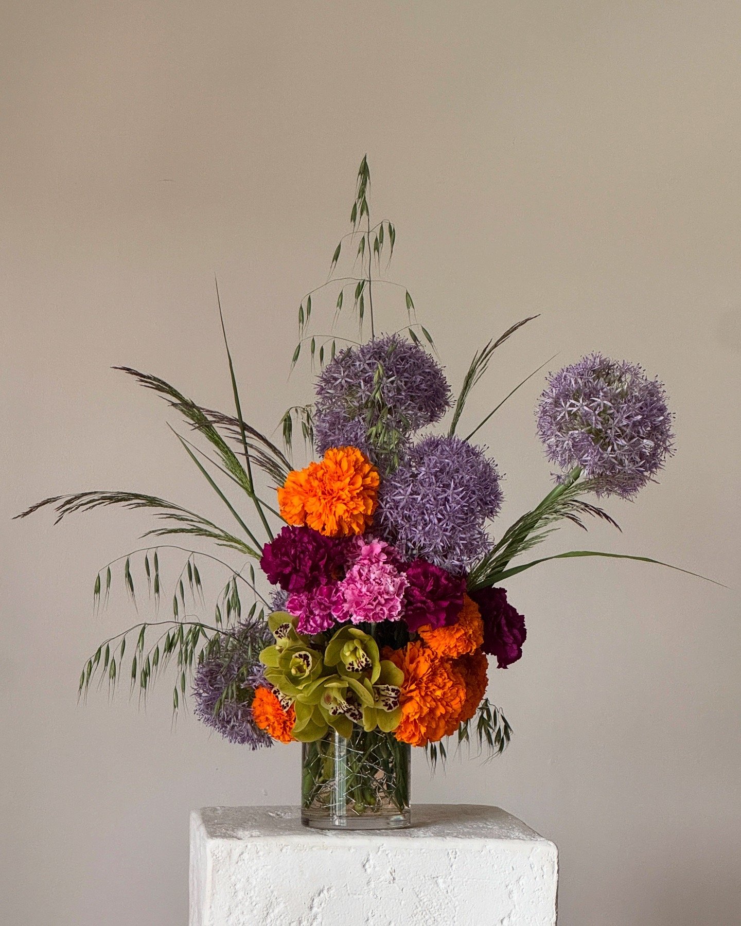 Colorful flower arrangement in a glass vase on a white pedestal against a plain light-colored wall.
