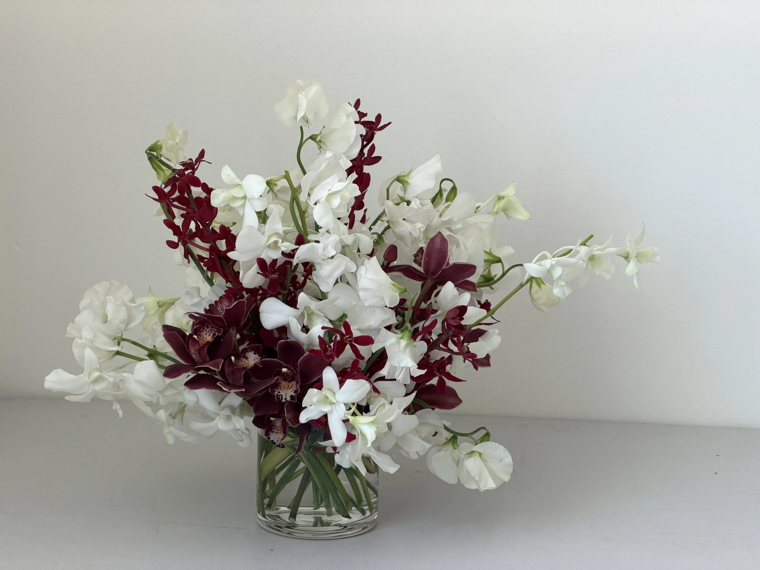 A clear glass vase containing a bouquet of white and dark red flowers, placed against a plain white background.
