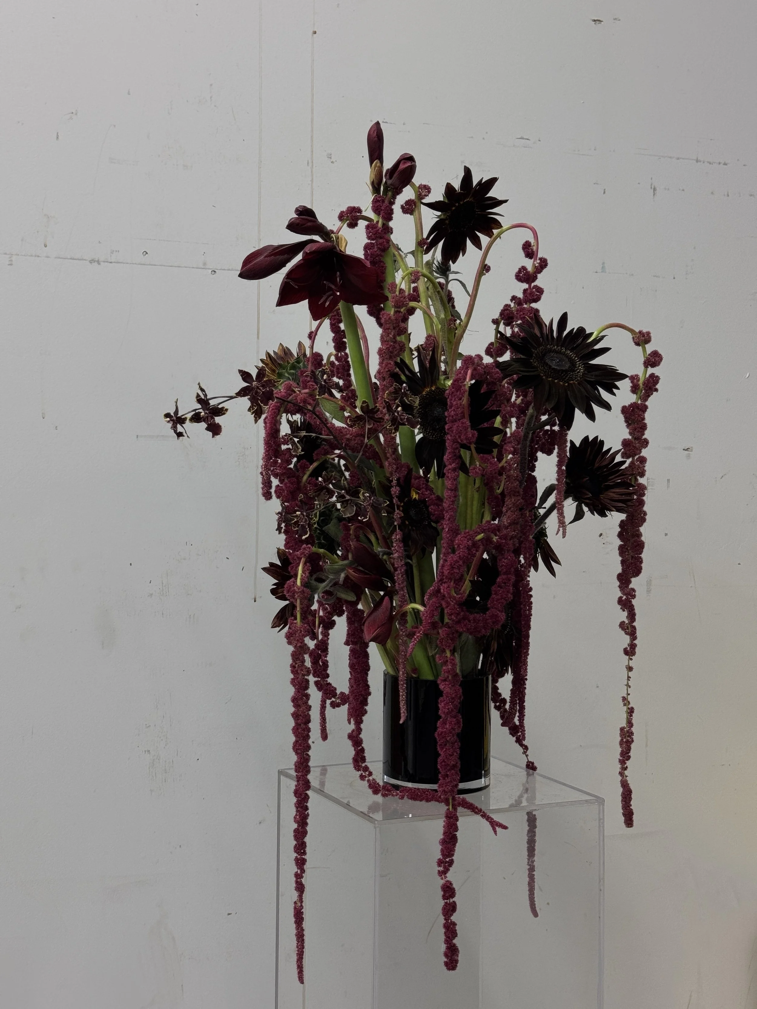An art installation of a tall black vase with artificial dark red and black flowers and long pinkish-red hanging strands, displayed against a plain light-colored wall.