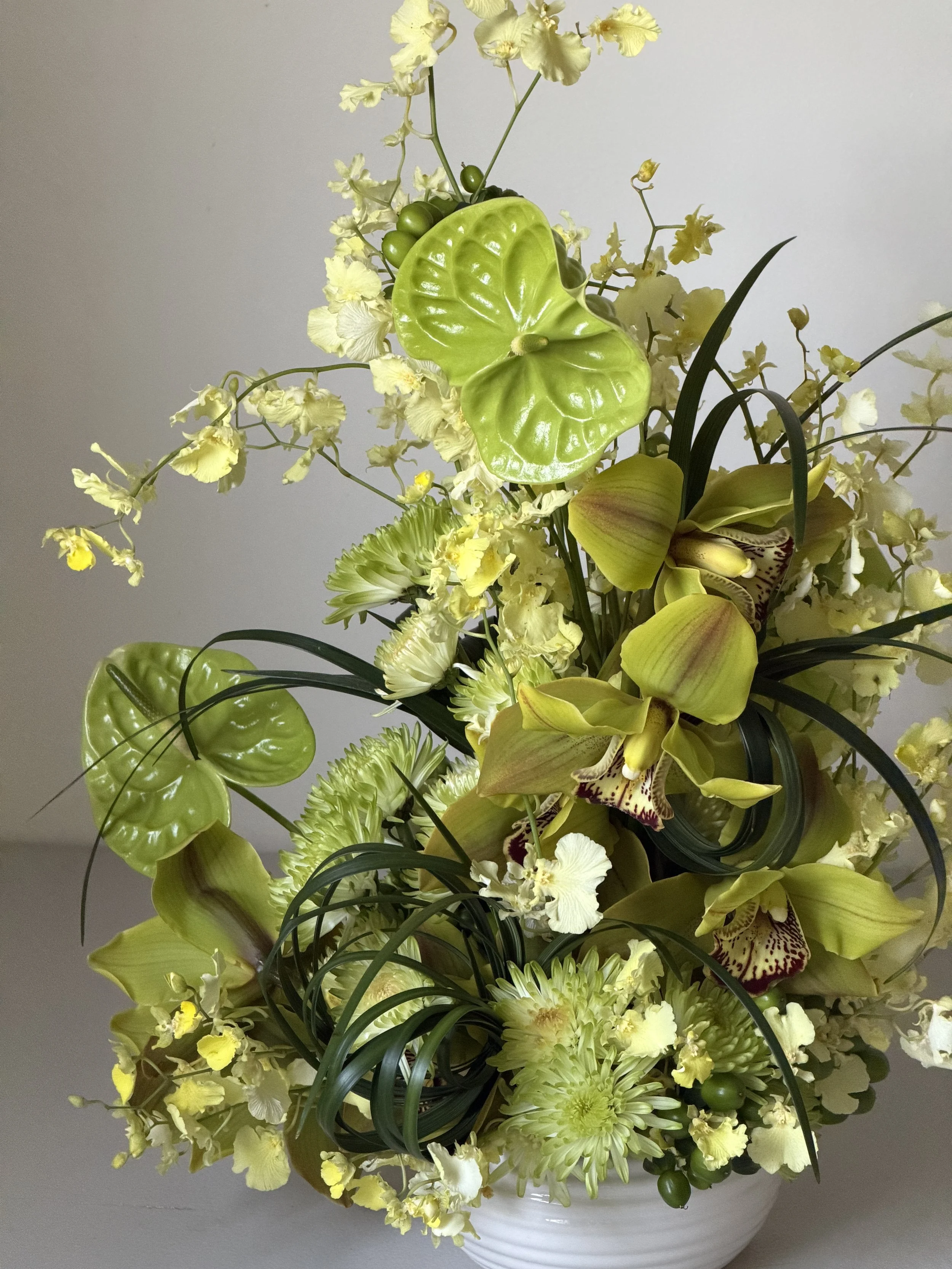 A floral arrangement with yellow and green flowers, including lilies, orchids, and anthuriums, in a white ceramic vase.