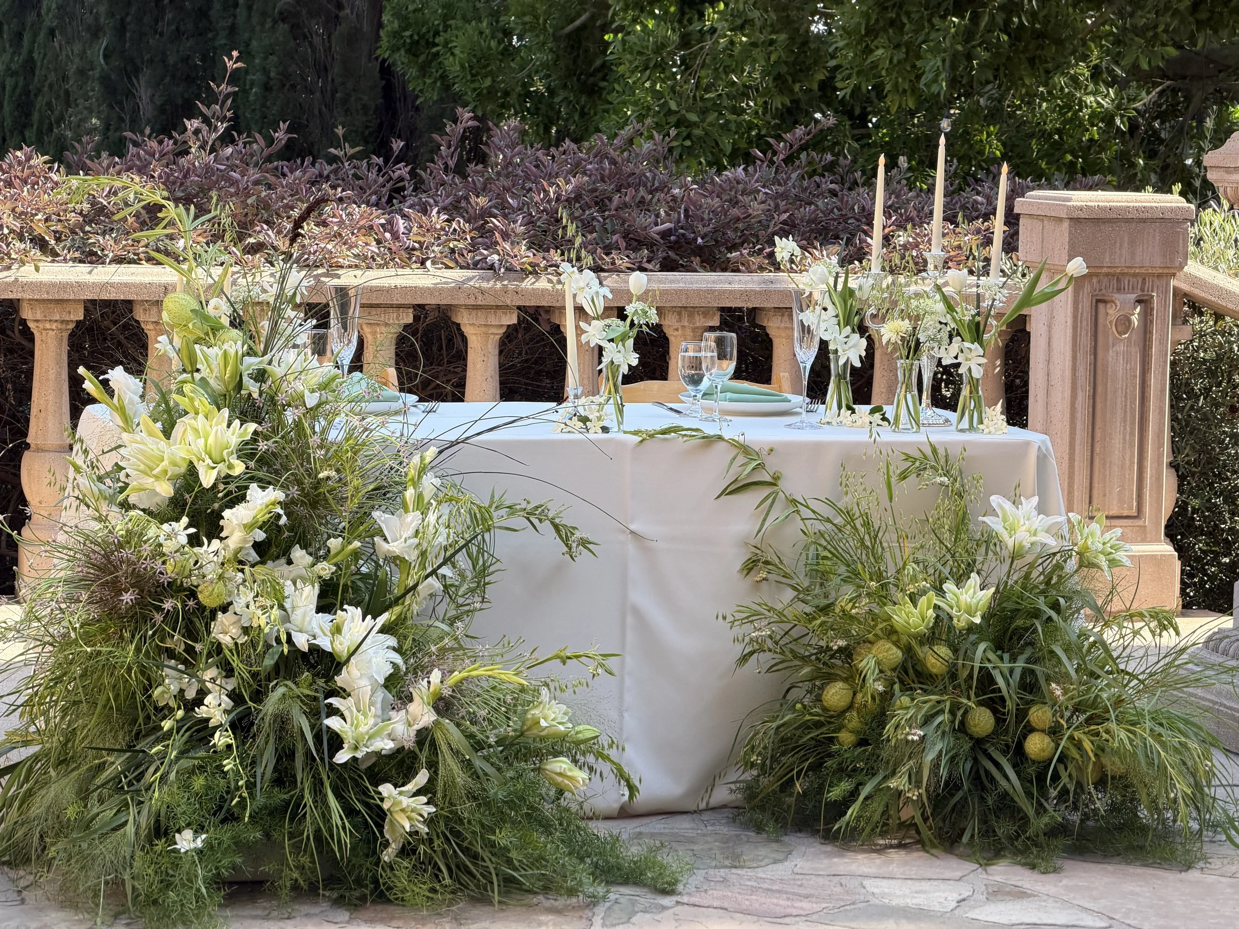 A dining table outdoors decorated with white flowers, greenery, tall candles, glassware, and tableware, set against a stone balustrade and lush green foliage.
