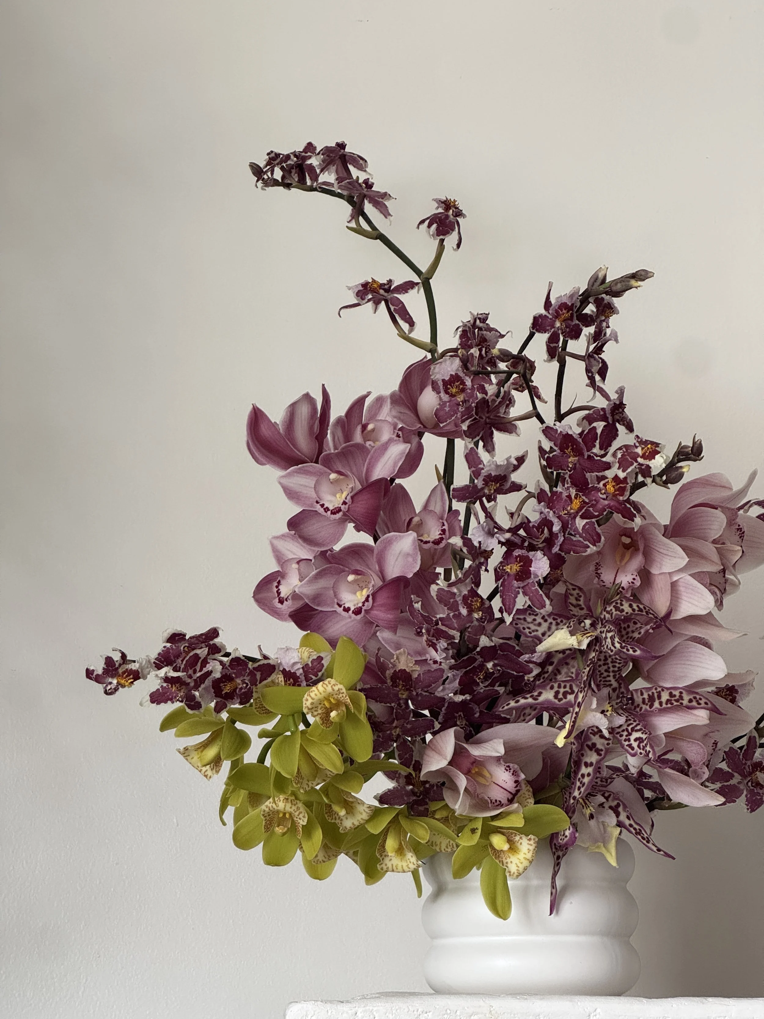 Elegant flower arrangement with pink, purple, and yellow orchids in a white vase against a plain white wall.