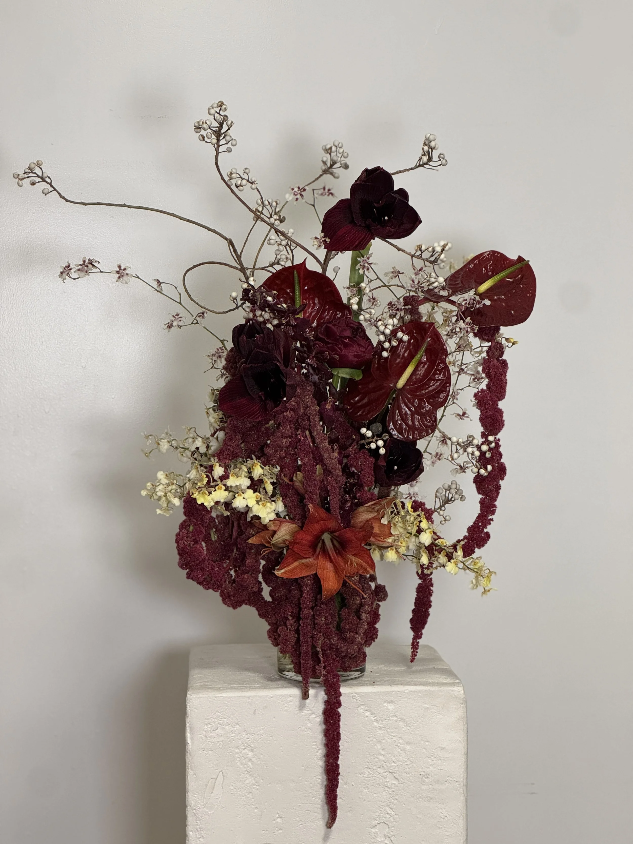 A tall floral arrangement in a clear glass vase features dark red flowers, white berries, and small pink flowers with thin, twisting branches, placed on a white pedestal against a light-colored wall.