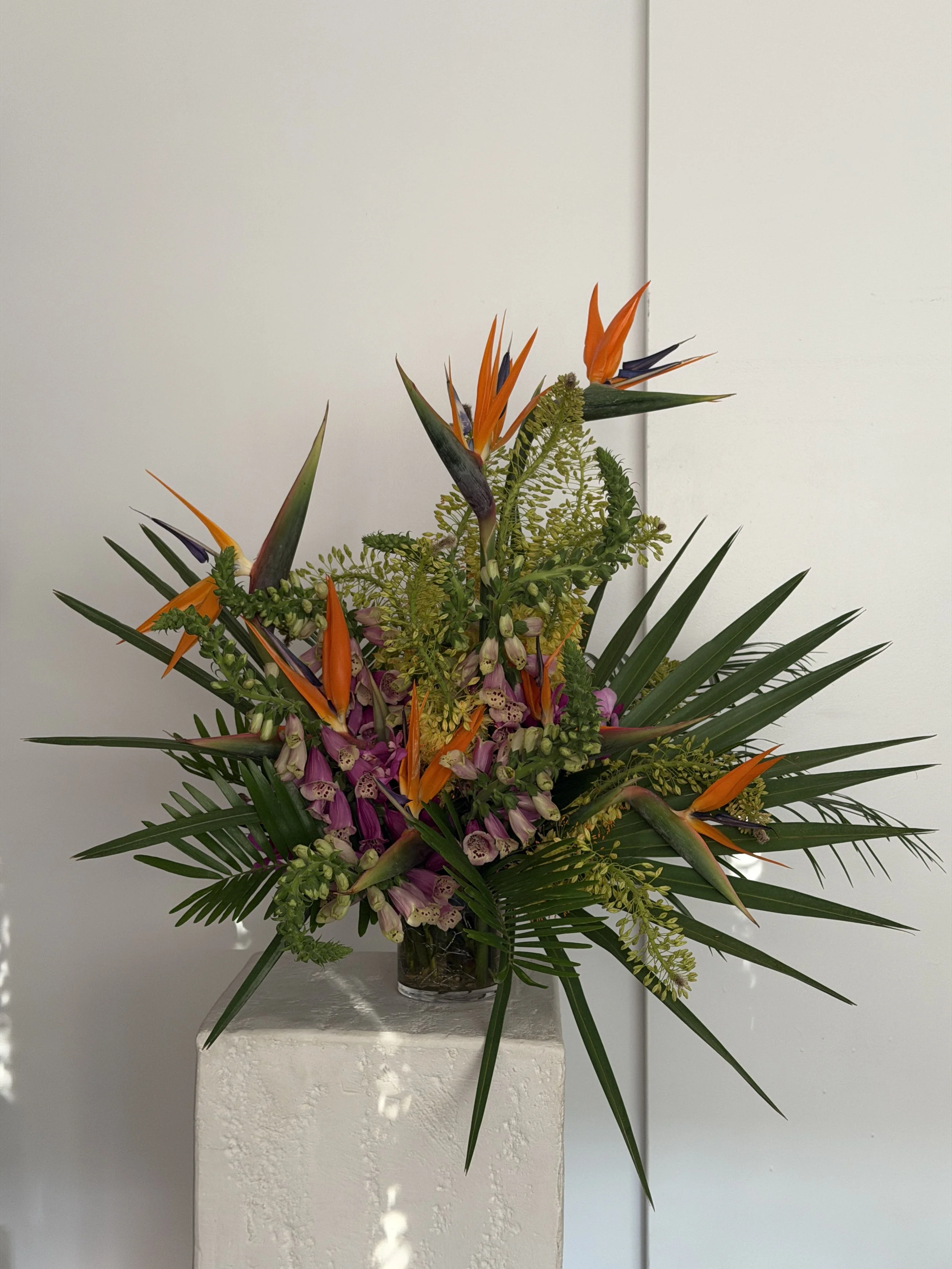 A tall floral arrangement with orange bird-of-paradise flowers, purple orchids, and greenery, placed in a glass vase on a white pedestal against a white wall.