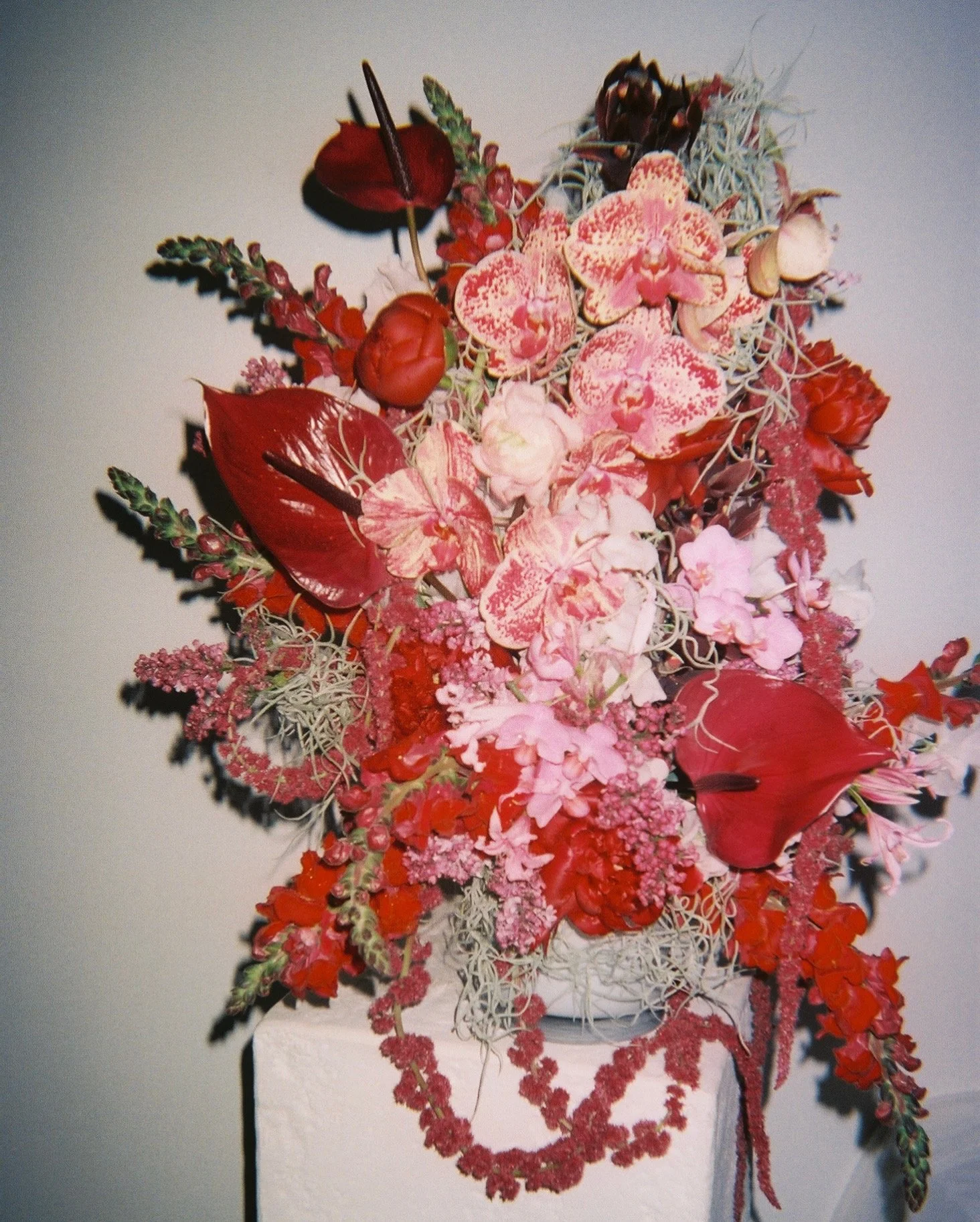 A colorful floral arrangement with pink, red, and green flowers, leaves, and accents displayed on a stand against a plain light background.
