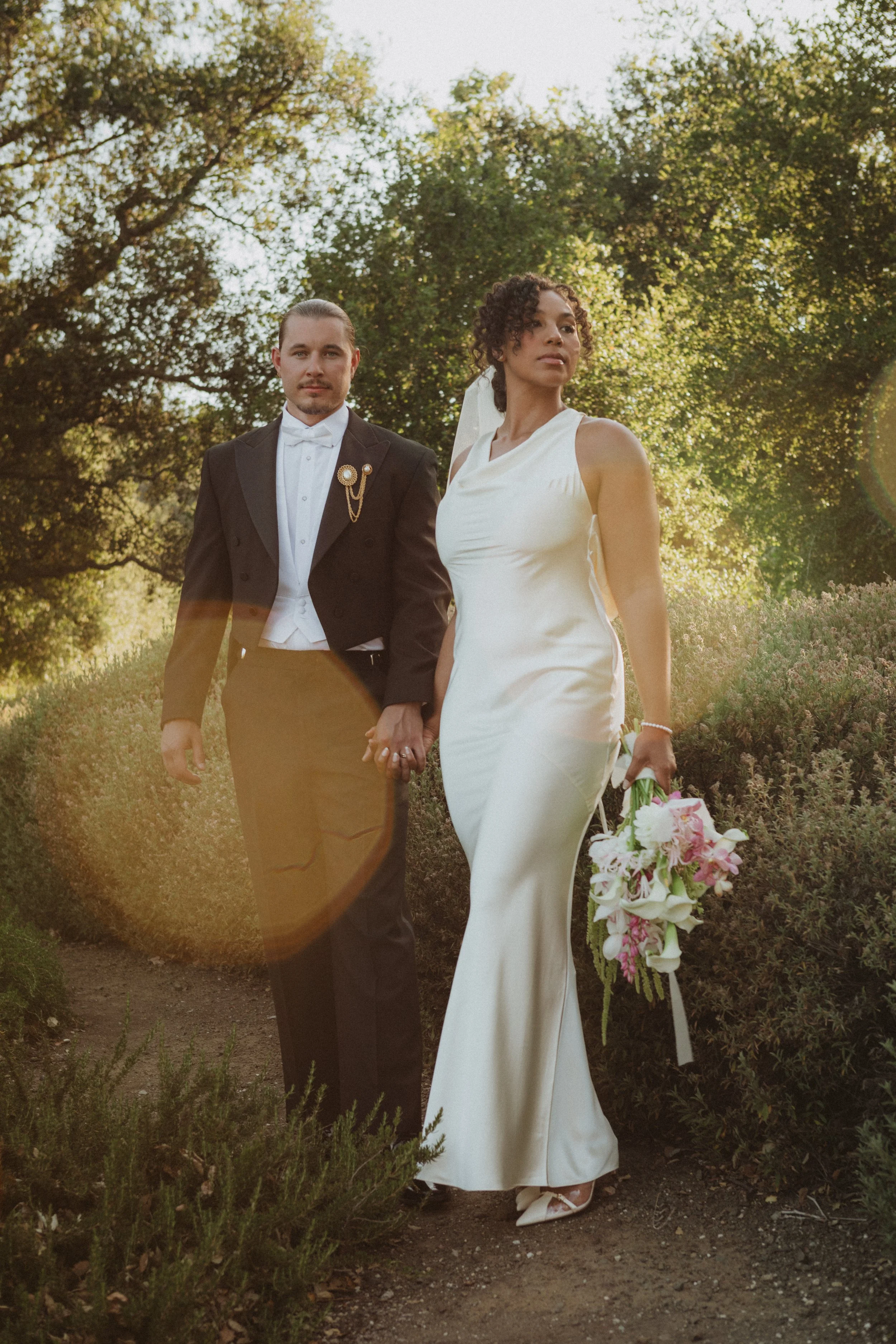 A couple dressed in wedding attire standing outdoors surrounded by trees and greenery.