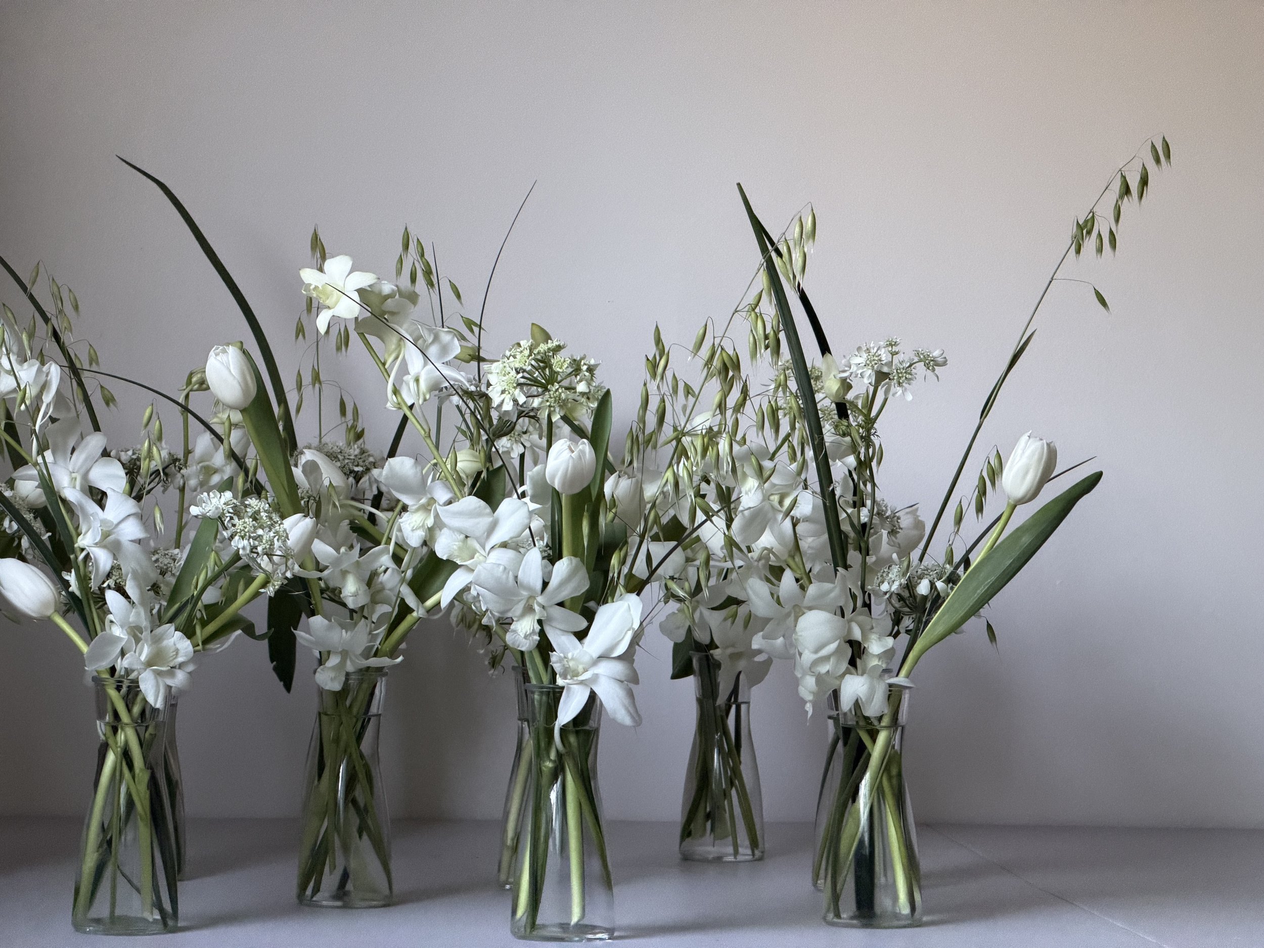 Four tall glass vases holding white flowers and green foliage, set against a plain background.