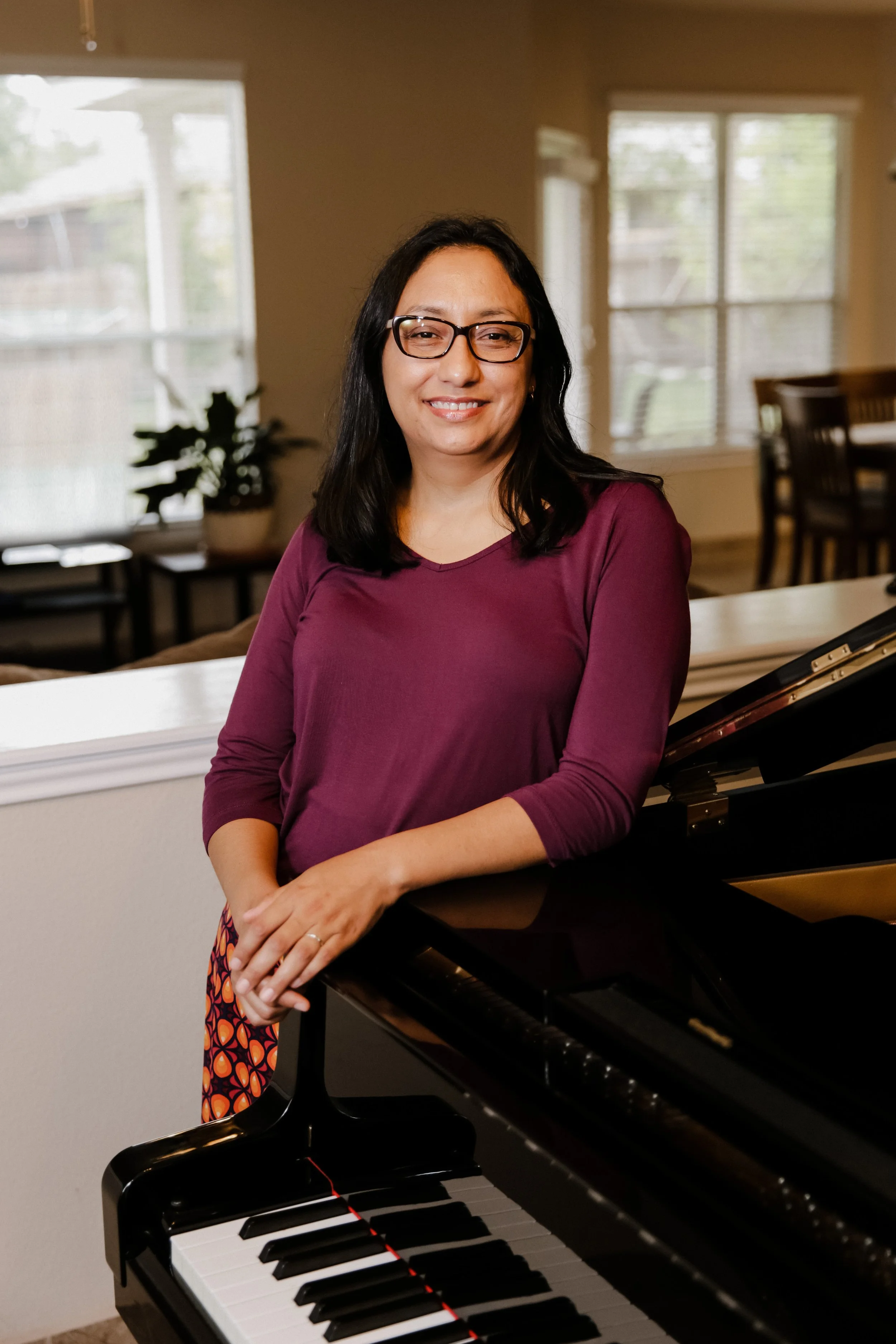 Mujer con gafas y blusa burdeos posando junto a un piano en una sala con ventanas grandes y plantas decorativas.
