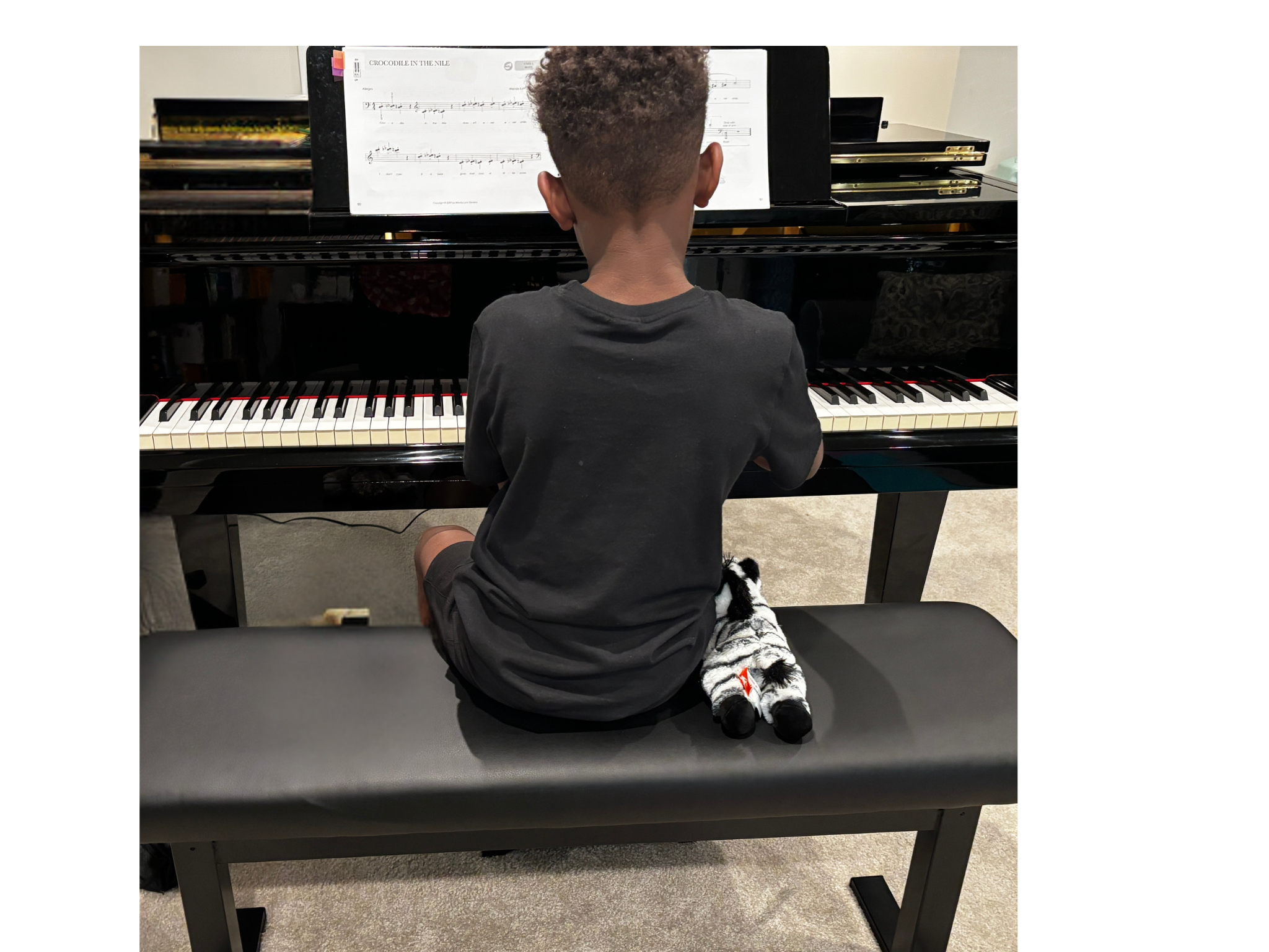 Niño tocando un piano negro en una sala de música, sentado en una banca. Tiene el cabello rizado y usa camiseta gris, con calcetines con patrones de tigre y unos zapatos negros y blancos.