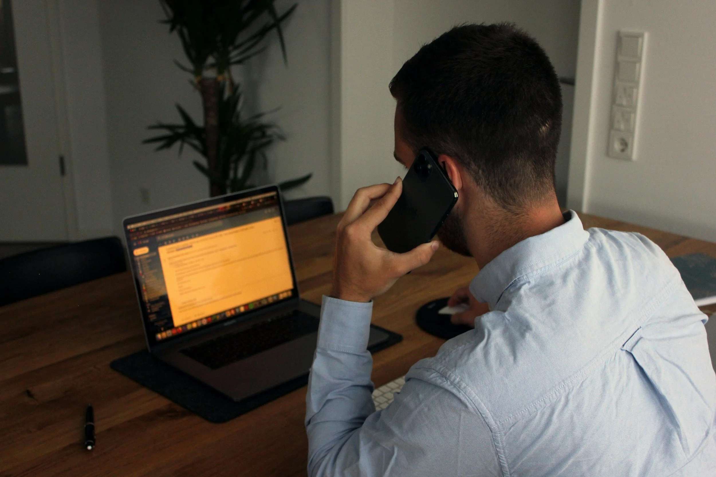 A man with short dark hair wearing a light blue shirt is talking on a black cell phone while sitting at a wooden desk. There is a laptop open in front of him displaying an email or document, a black pen, and a computer mouse on the desk. A large plant is in the background near a white wall with light switches.