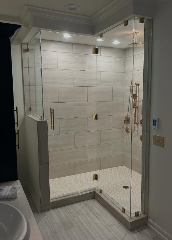 A glass shower enclosure with brass hinges and handles in a bathroom, featuring beige tiled walls and a white hexagonal tile floor inside the shower, with a rainfall showerhead and handheld shower attached to the wall.
