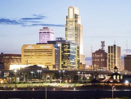 Nighttime cityscape of downtown with tall buildings, including a prominent skyscraper, illuminated windows, and a park in the foreground.