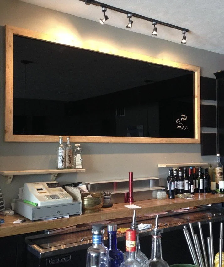 A large wall mirror with a wooden frame hangs above a bar counter displaying bottles of alcohol, a cash register, and bar tools.